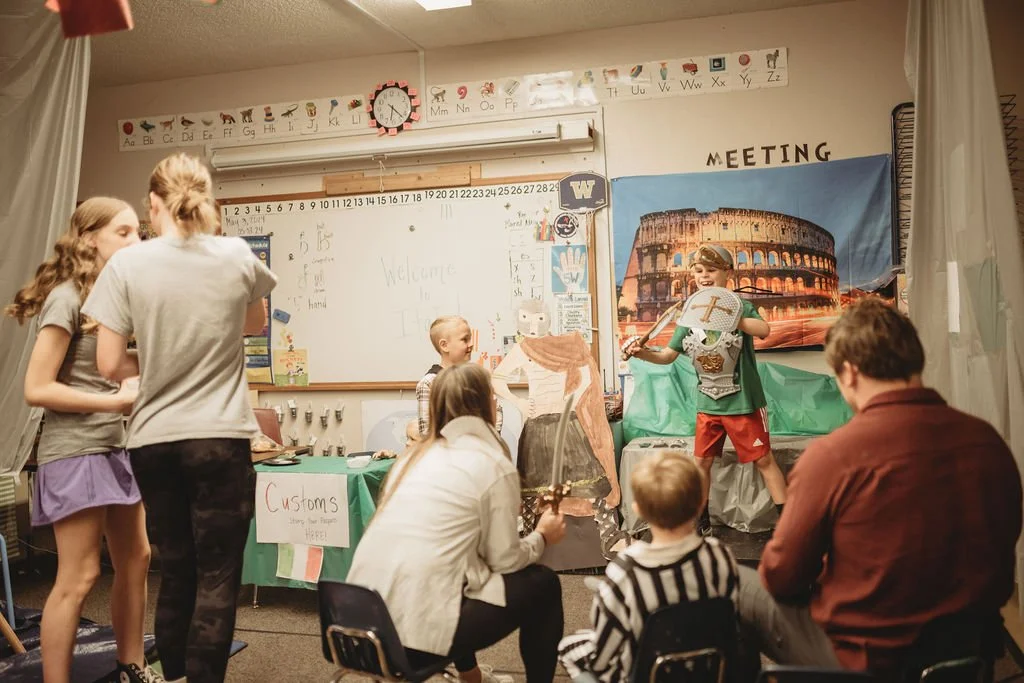Children and adults in a classroom with a puppet show setup, including a painted backdrop of the Colosseum, and a whiteboard with 'Welcome' written on it.