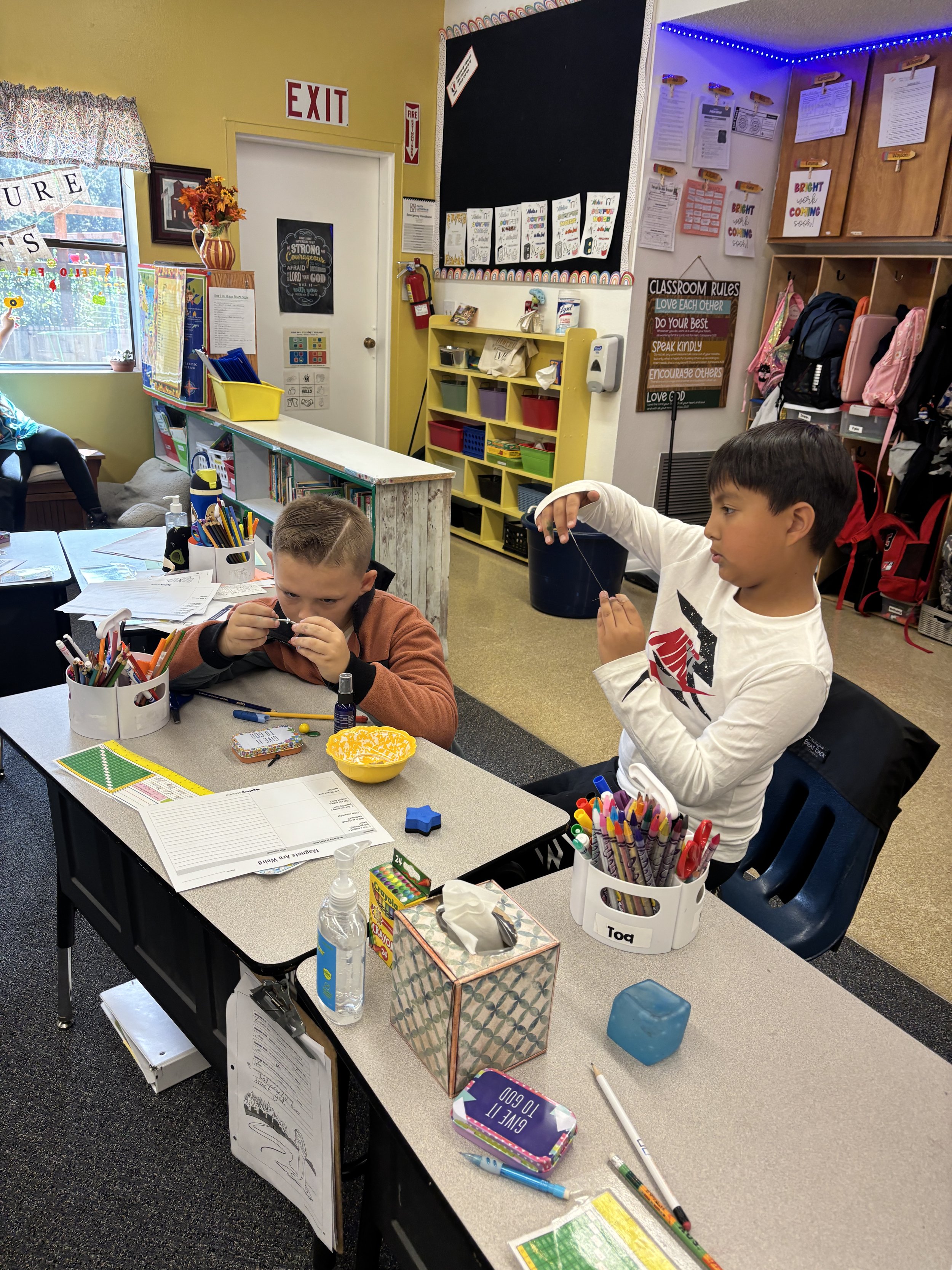 Two young boys are sitting at a school desk. The boy on the left is examining a small object closely, while the boy on the right is holding a string or thread. The desk is cluttered with school supplies, including pens, markers, a bottle of hand sanitizer, and a tissue box. In the background, there is a yellow bookshelf filled with baskets, a black bulletin board with papers, a door with an EXIT sign, and a window with decorative curtains. The classroom has signs and educational posters on the walls.