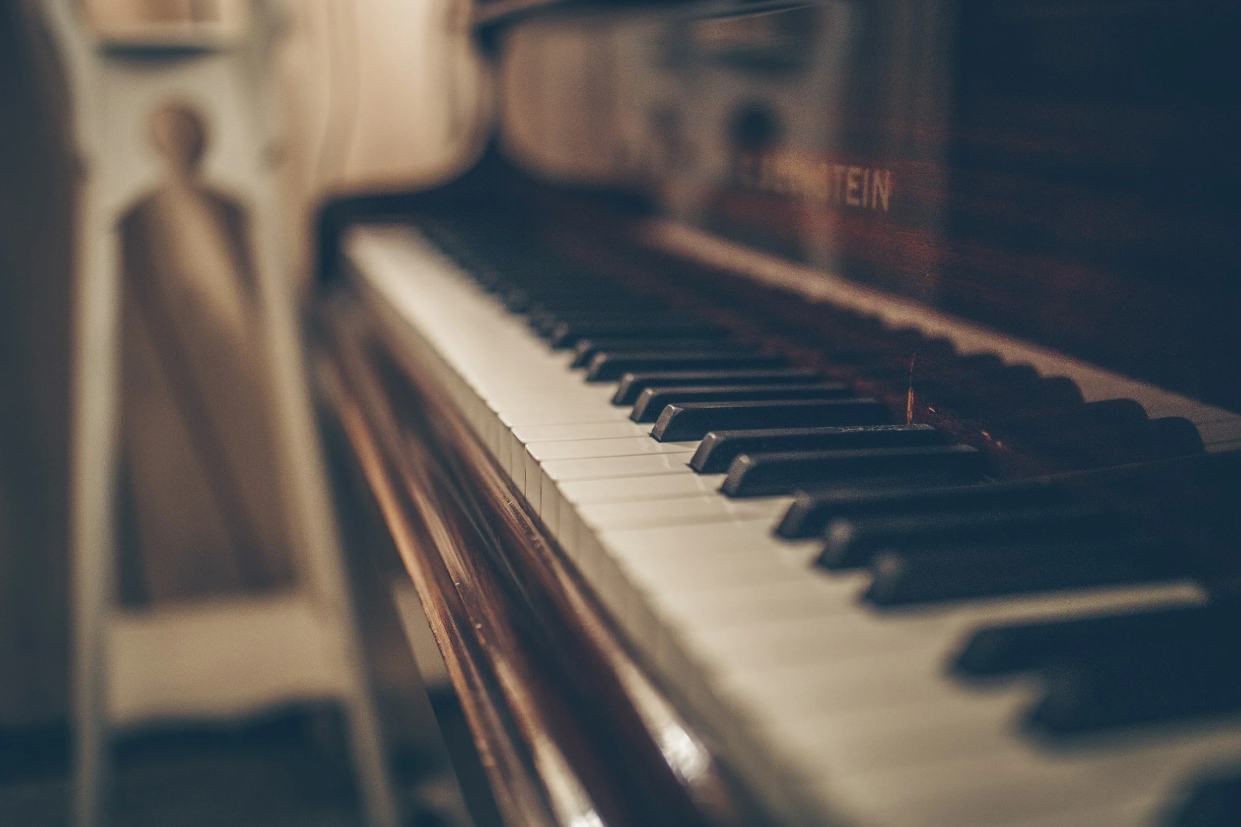 Close-up of a wooden piano keyboard with black and white keys, with a blurred chair in the background.