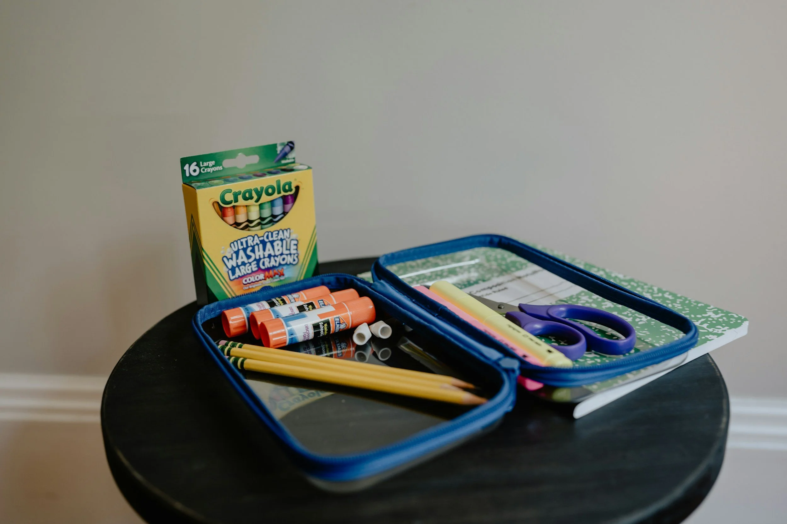 A small black round table holding school supplies: a box of Crayola washable large crayons, a blue pencil case with yellow pencils, glue sticks, scissors, a green notebook, a yellow highlighter, and a pink highlighter.