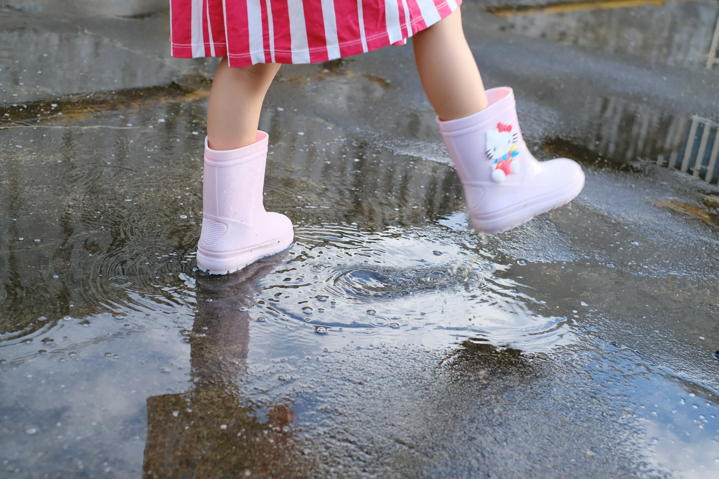 A young girl wearing pink rain boots with Hello Kitty design, standing on a wet concrete surface, splashing in a puddle.