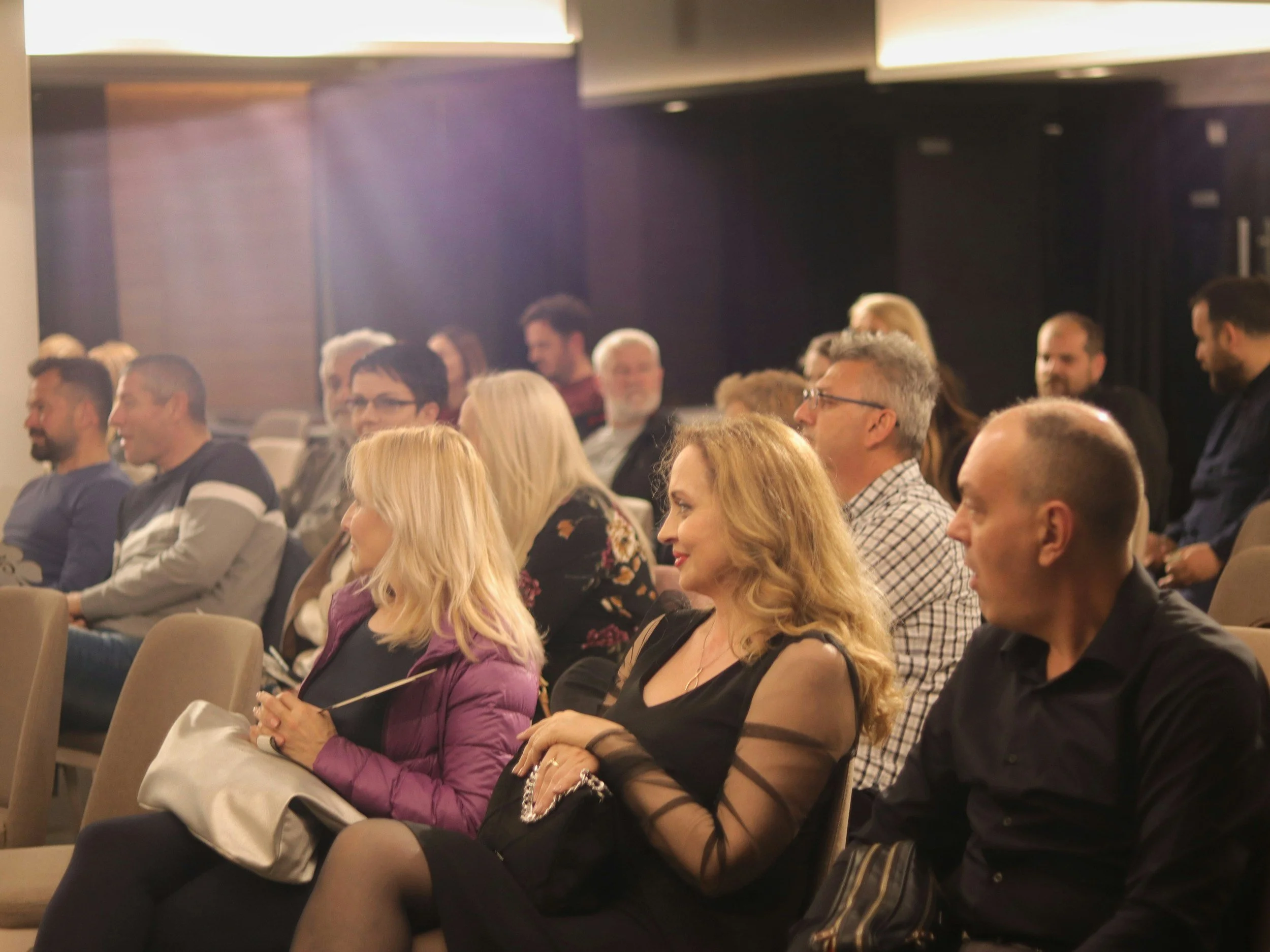 Audience members seated and smiling at a conference or presentation, with a woman in the foreground wearing a black dress with sheer sleeves.