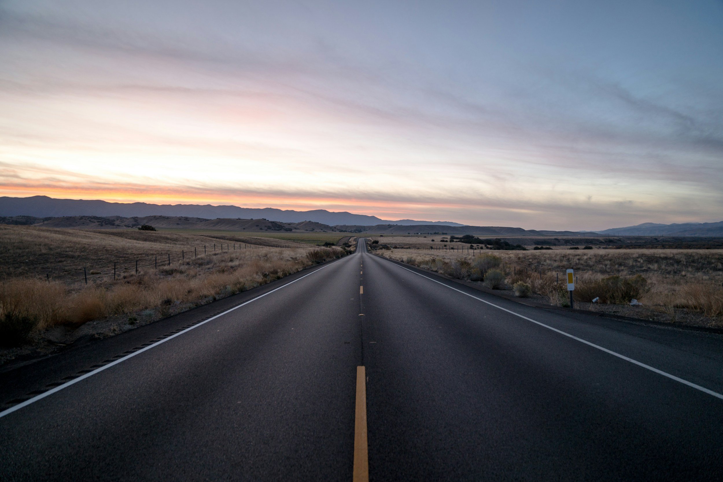 Empty open road stretching into the distance during sunset with mountains in the background.