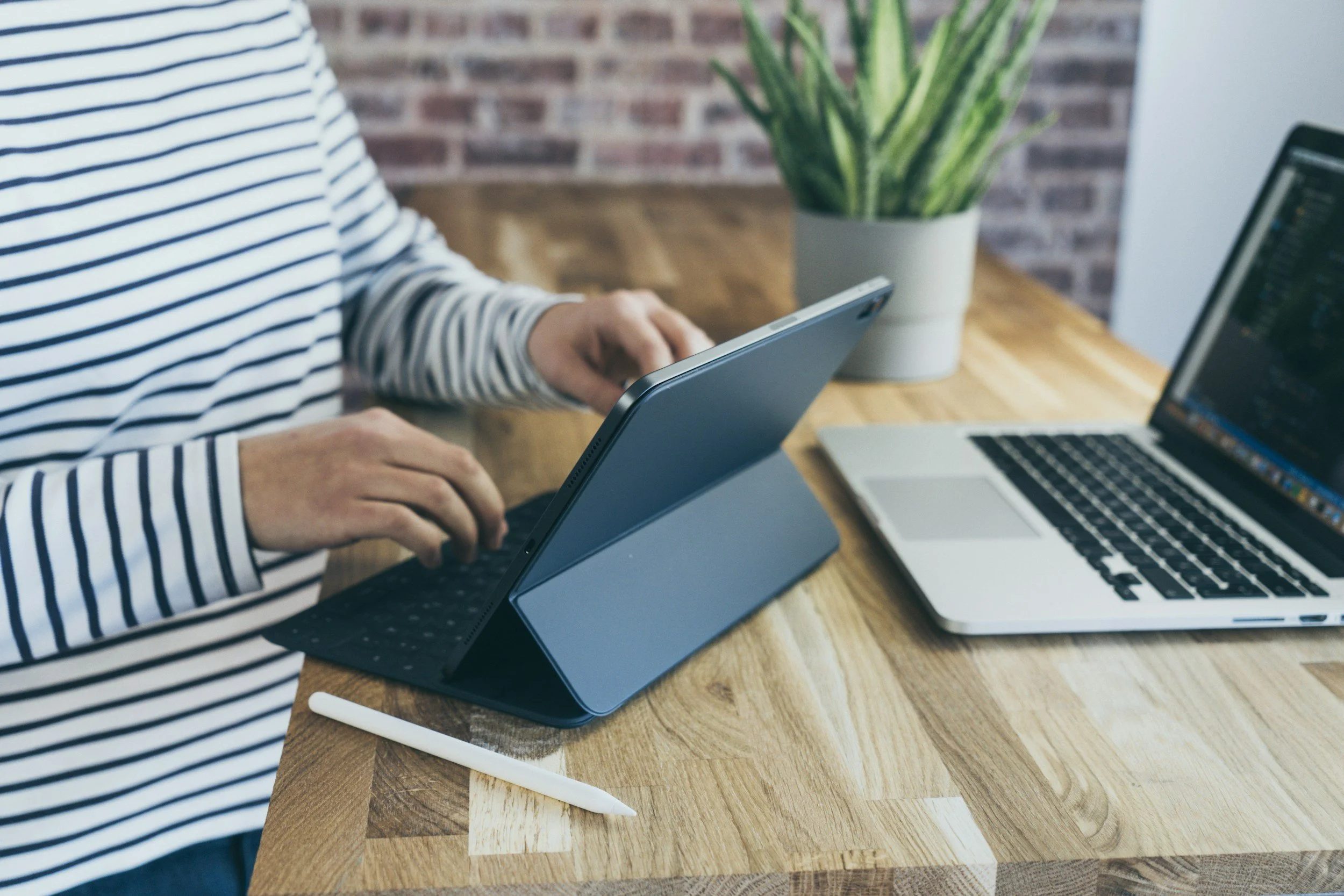 Person in a striped shirt using a tablet with a keyboard case on a wooden desk. There is an open laptop, a white stylus, and a potted plant on the desk, with a brick wall in the background.