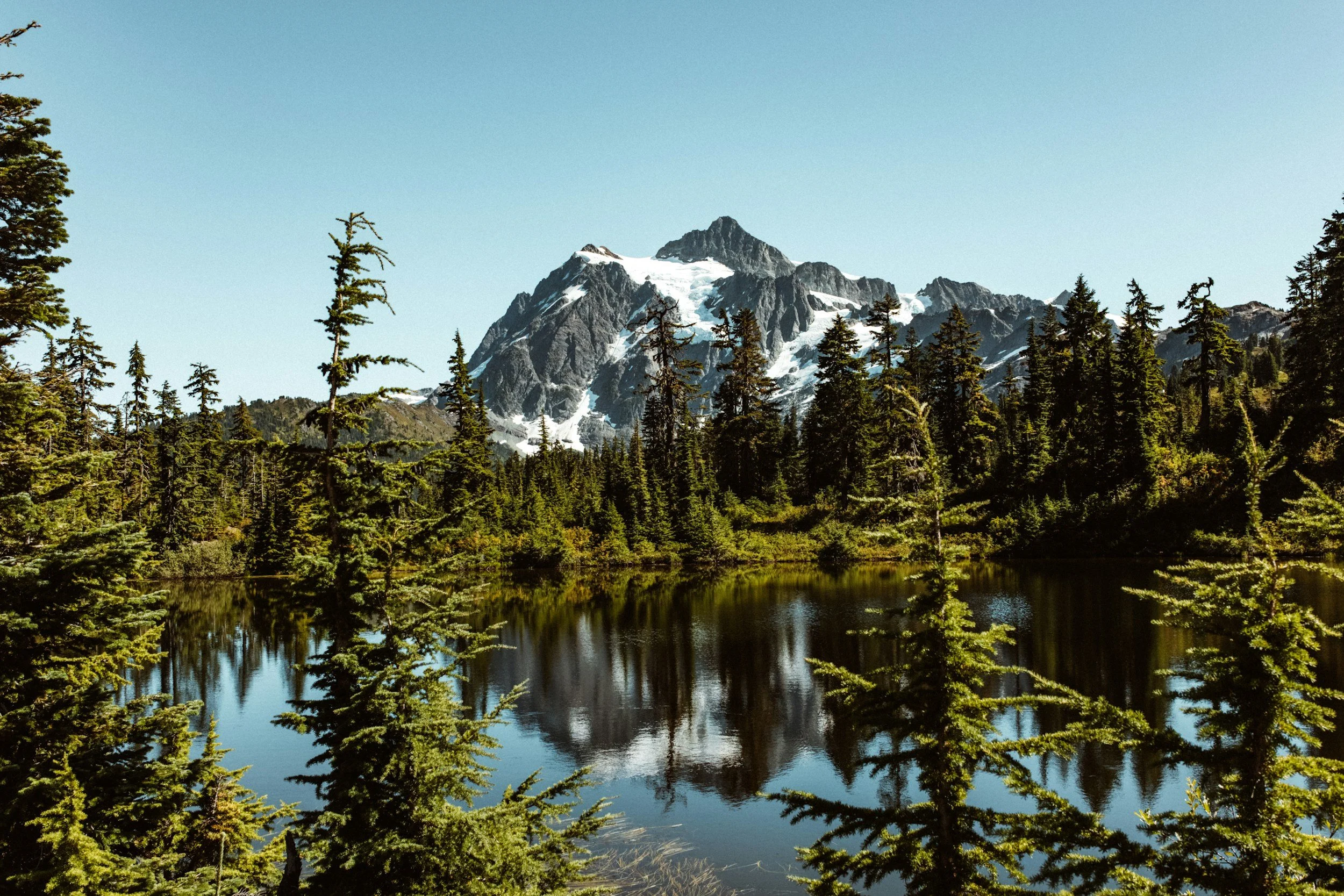 A mountain with snow patches, surrounded by forest, reflected in a lake.