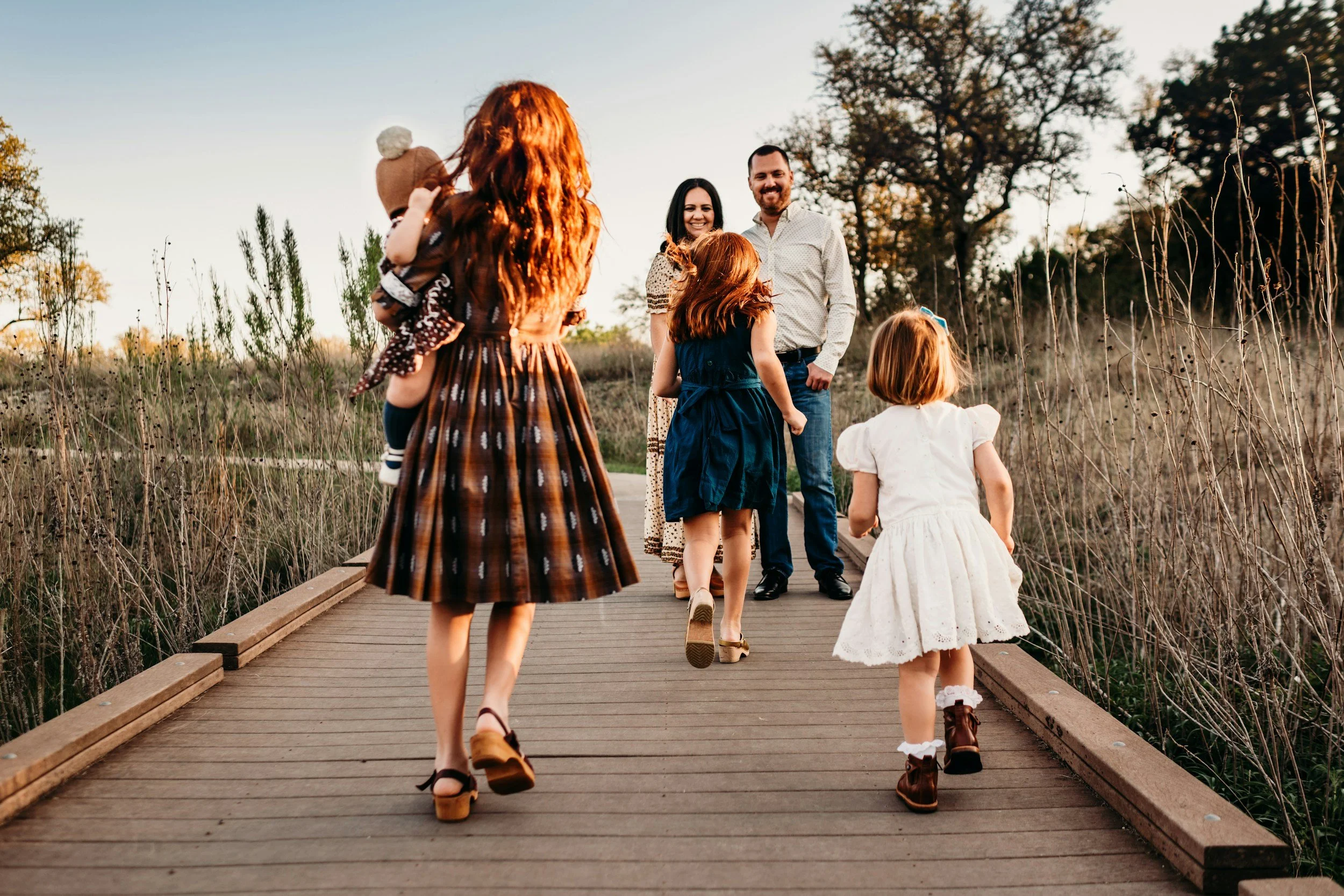 A group of children and two adults smiling and walking towards the camera on a wooden bridge outdoors during sunset.