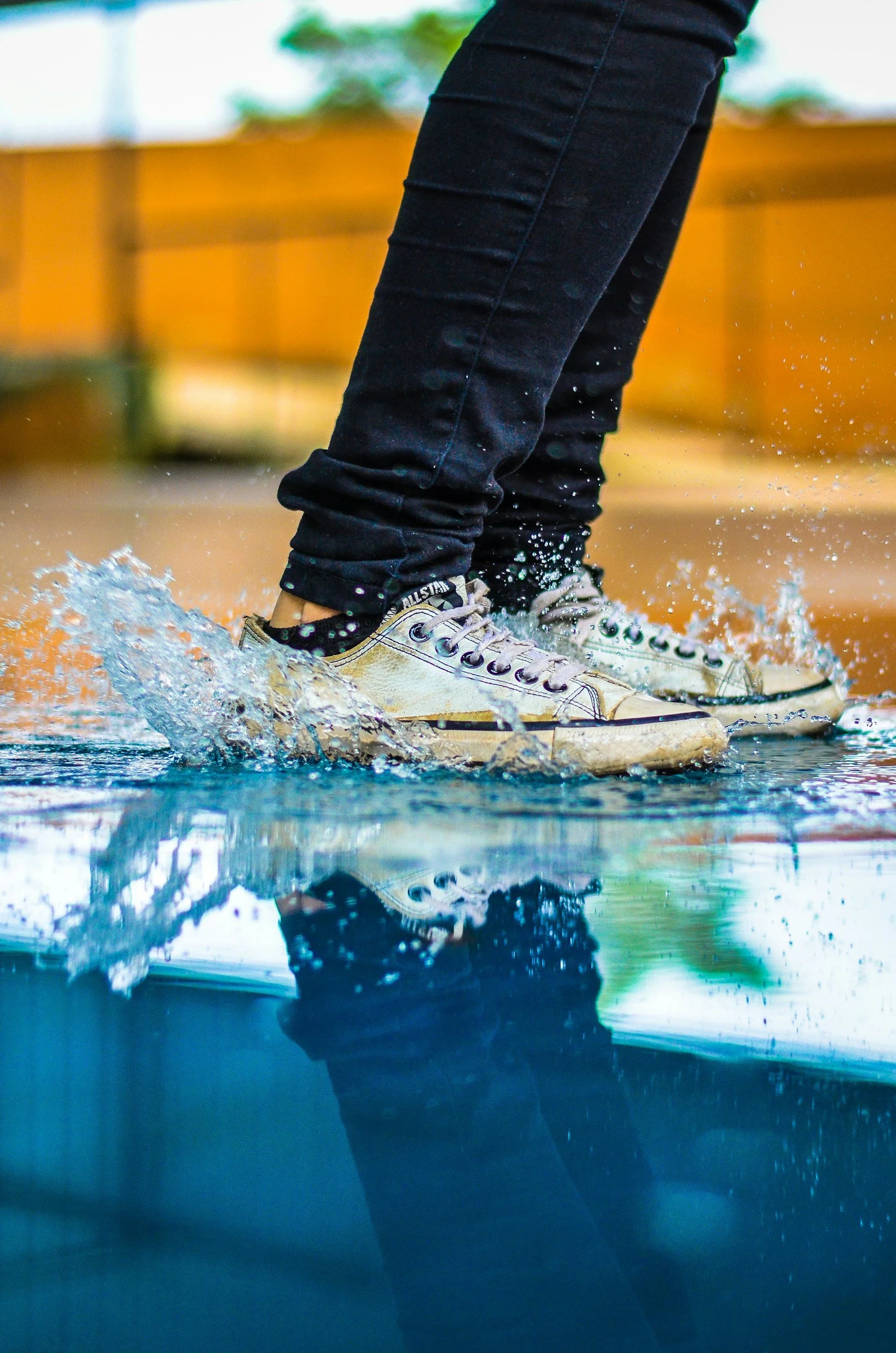 Person wearing black pants and white sneakers walking through a puddle of water, splashing water around.