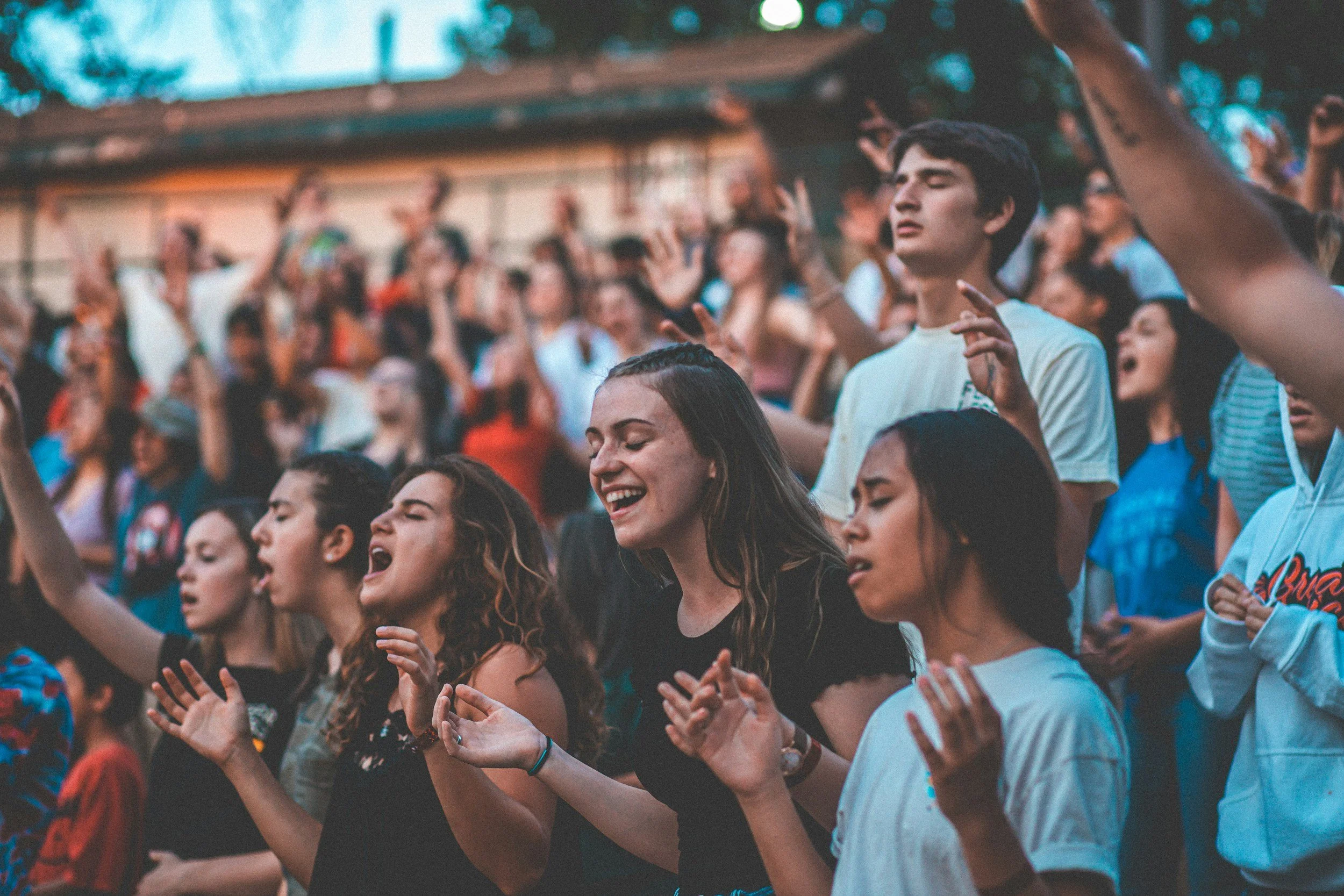 People at a concert or outdoor event with many individuals singing, dancing, and enjoying music, with some raising their hands and eyes closed.