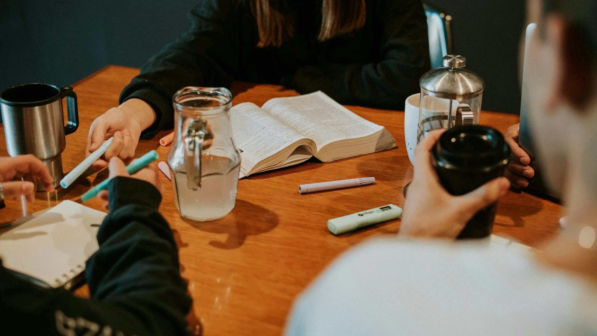 Group of people sitting around a wooden table with coffee cups, a water pitcher, an open book, markers, a notebook, and a reusable coffee cup, engaging in a discussion.