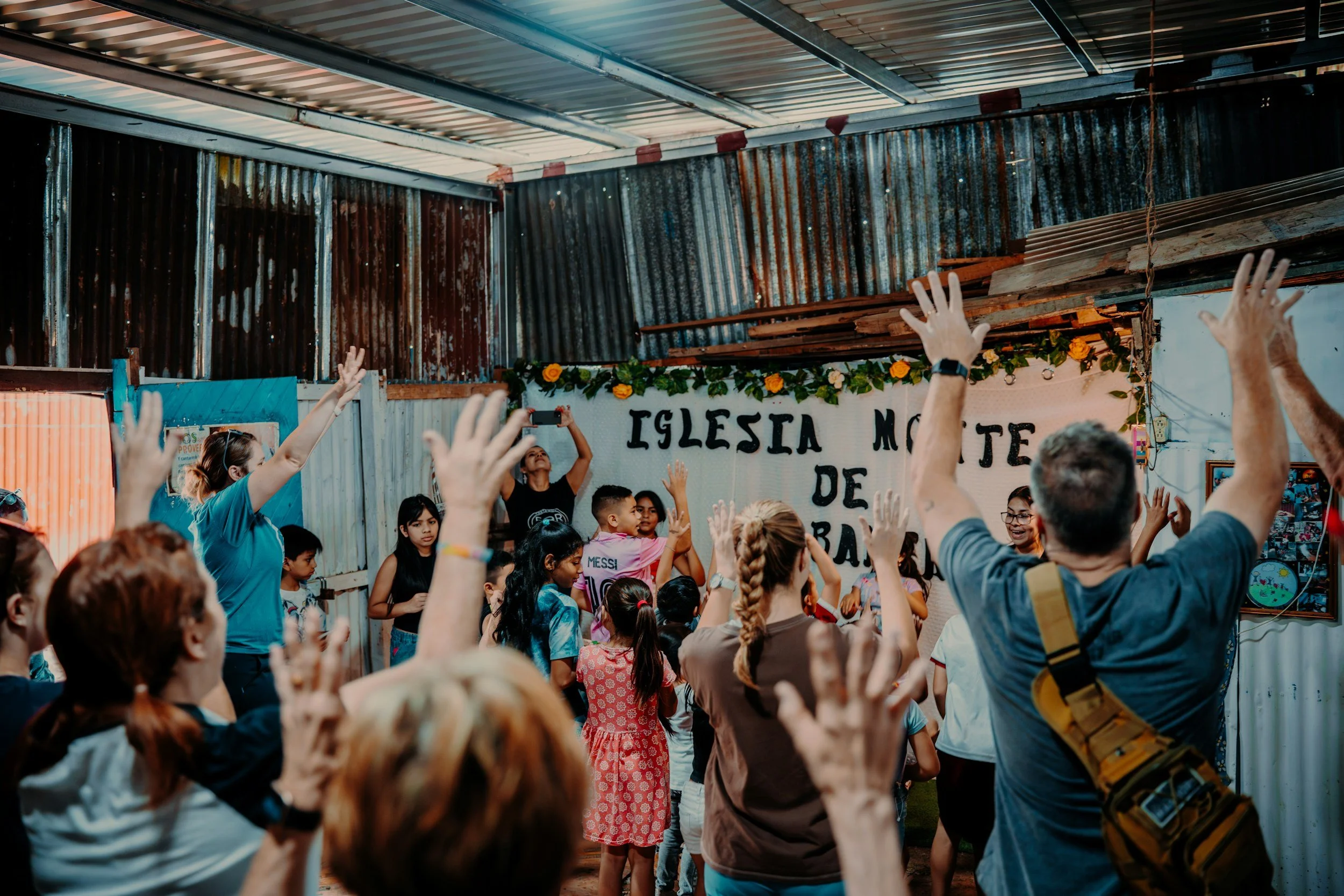 People dancing and raising their hands in a celebration inside a rustic building with corrugated metal walls. A banner in the background reads 'IGLESIA M.'.