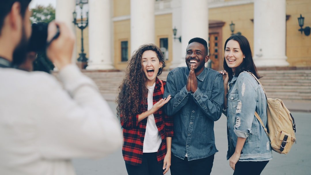 A group of three friends taking a photo, standing in front of a historic building with columns, stairs, and vintage street lamps. The friends are smiling and appear happy.