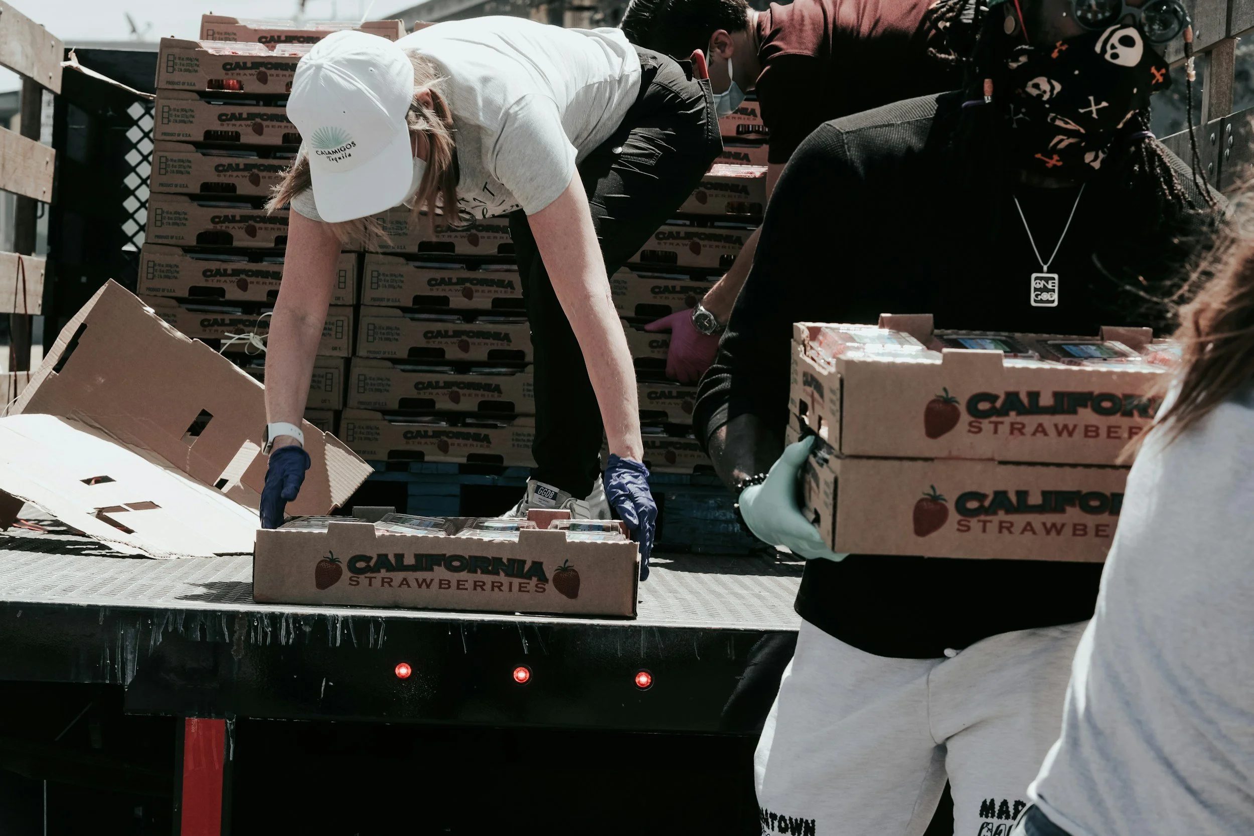 People working outdoors handling cardboard boxes of strawberries labeled 'California Strawberries' at a produce distribution event.