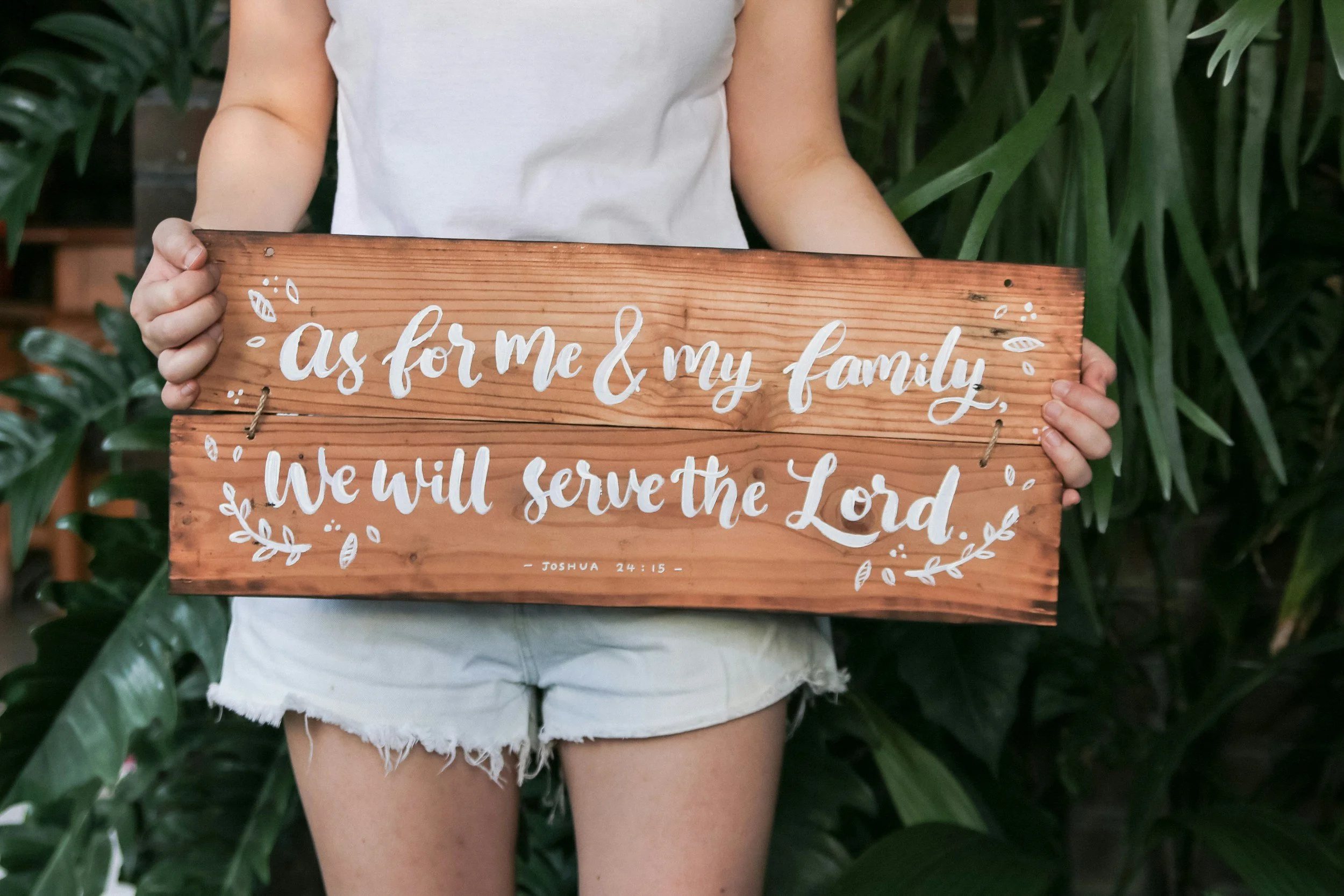 Person holding a wooden sign with the words "as for me & my family, we will serve the Lord" from Joshua 24:15, with greenery in the background.