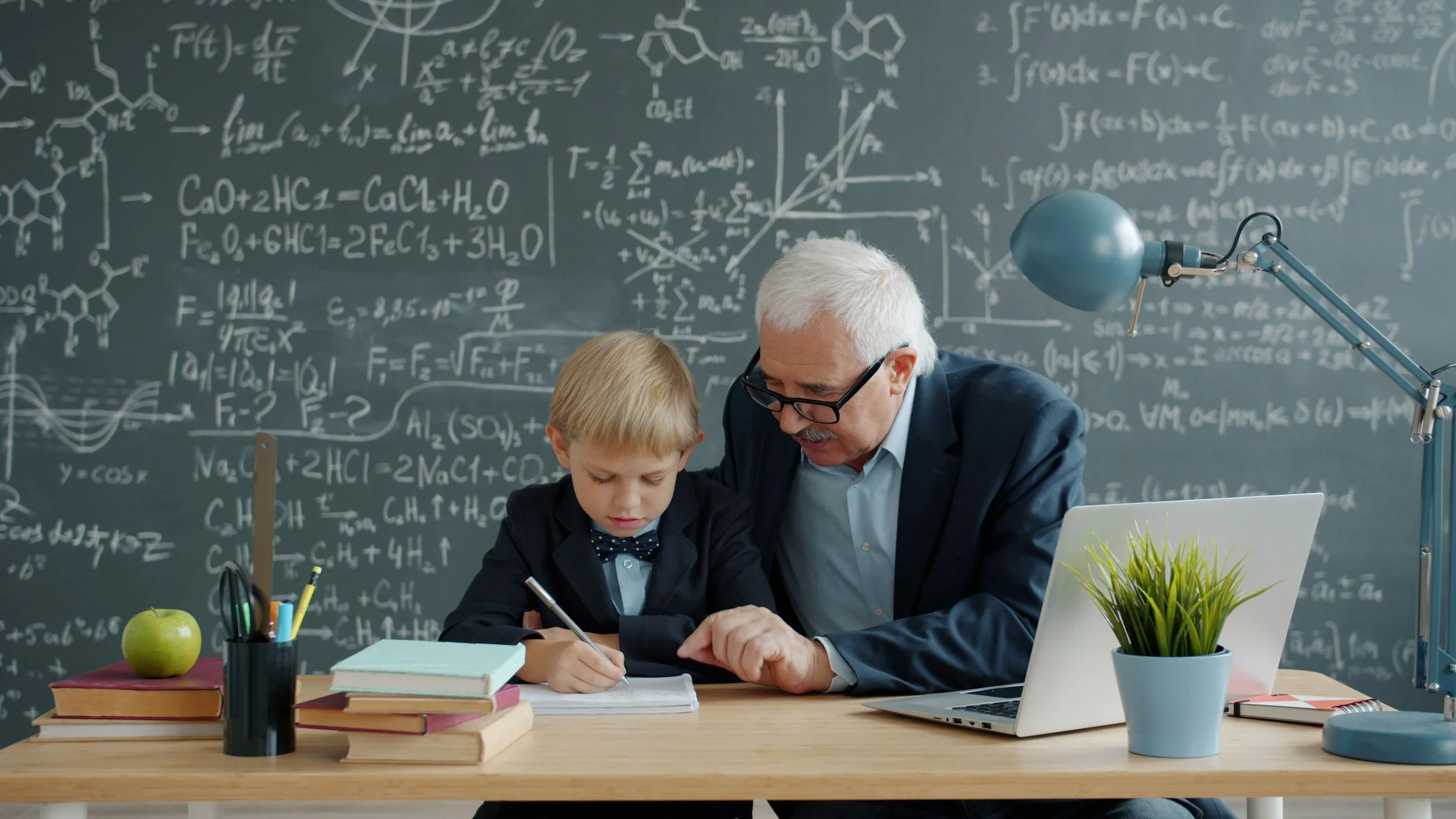 An elderly man and young boy sitting at a desk in a science classroom with a chalkboard filled with math and science diagrams behind them. The boy is writing in a notebook while the man guides him. The desk has books, a green apple, a plant, a laptop, and stationery.