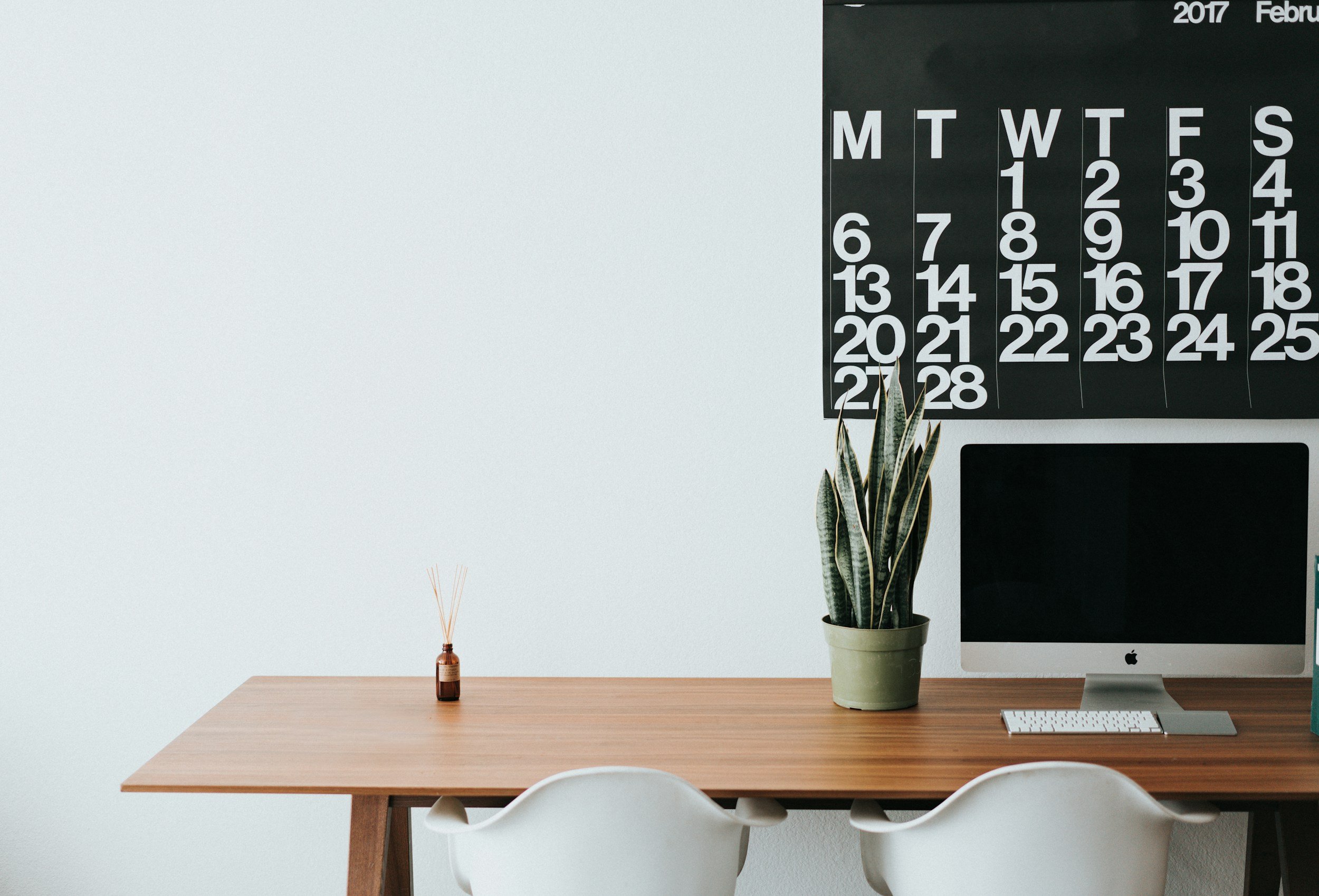 A minimalist workspace with a wooden desk, white chairs, a green potted snake plant, and a computer monitor. A black calendar with white numbers and days is on the wall.