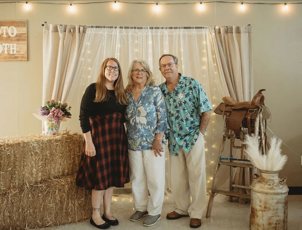 Three people standing together in front of a decorated backdrop with string lights, beige curtains, and a hay bale table with a flower arrangement. The left person is a young woman with long red hair, glasses, wearing a black top and a red plaid skirt. The middle person is an older woman with blonde hair, glasses, wearing a blue floral blouse and white pants. The right person is an older man with gray hair, wearing glasses, a blue floral shirt, and white pants.