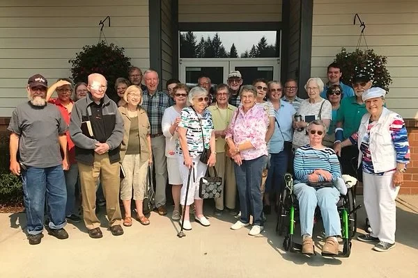 Group of elderly people and a few younger individuals standing outside in front of a building with two hanging flower baskets, smiling for the photo.