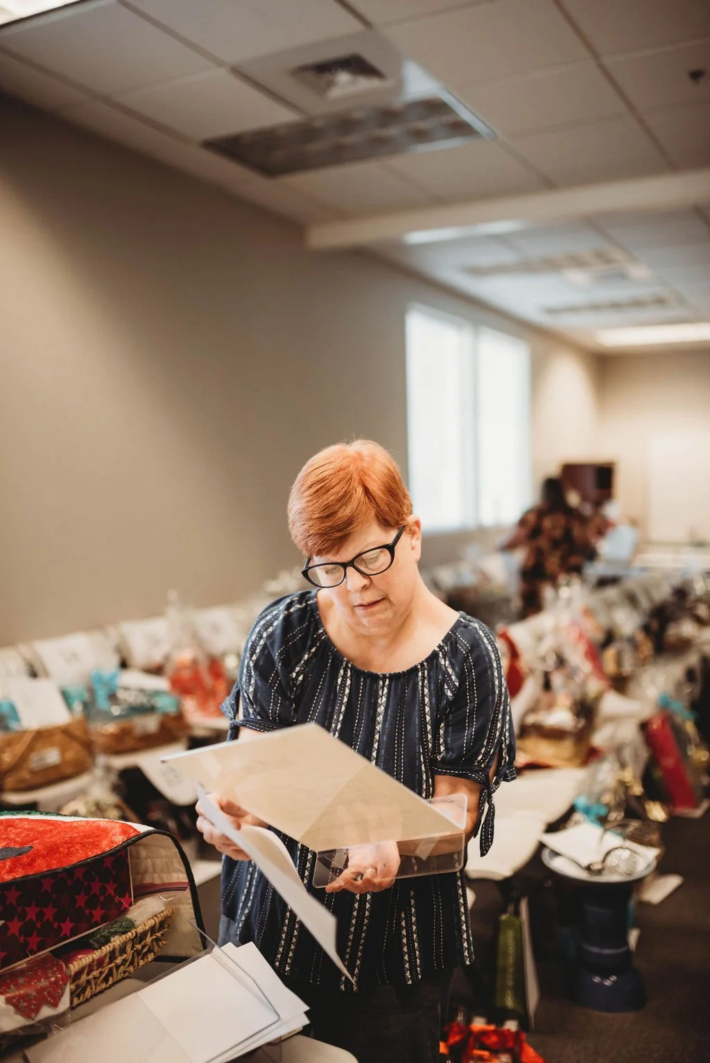 A woman with short red hair and glasses looking at a photo album in a room filled with tables of gifts and items, possibly at a gift exchange or holiday event.