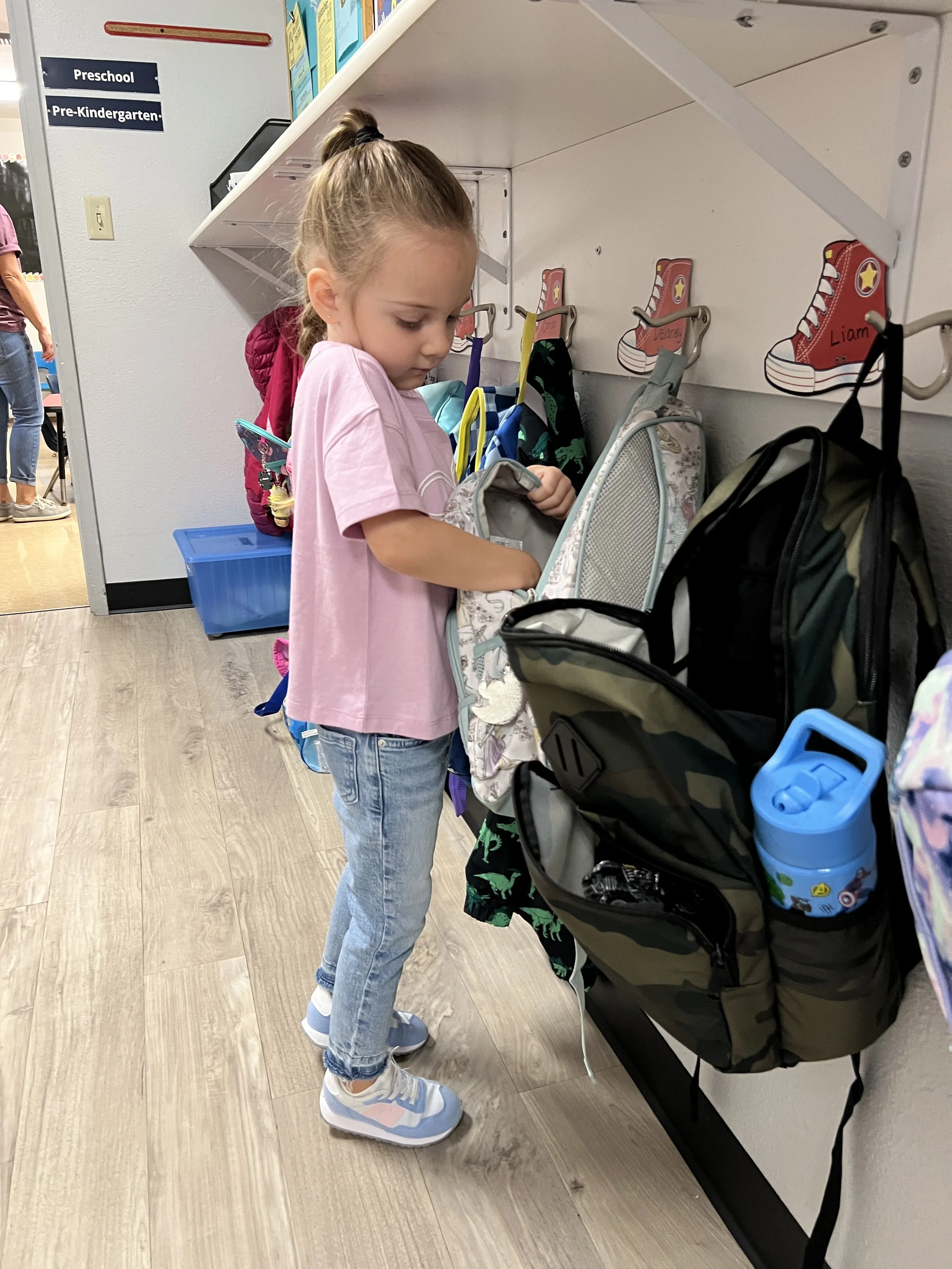 A young girl in a pink shirt and jeans stands at a coat hook rack in a preschool classroom, looking into her backpack. There are labeled signs for 'Preschool' and 'Pre-Kindergarten' on the wall, and decorative drawings of red sneakers with names inside.