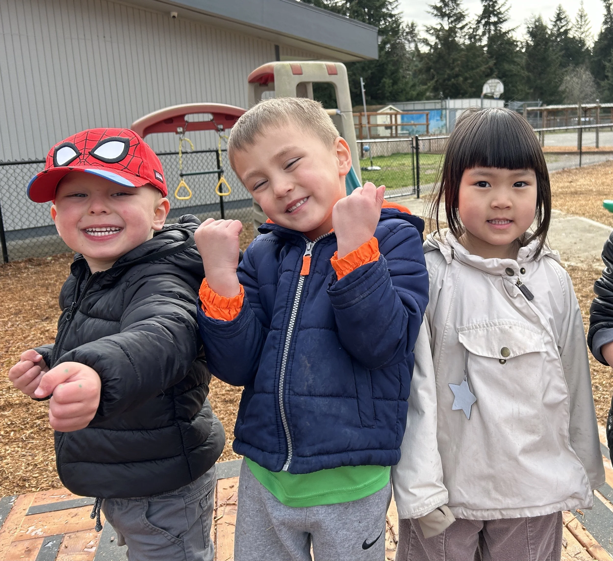 Three children standing outdoors, smiling and posing with their arms raised. The background includes playground equipment, a fence, and trees.