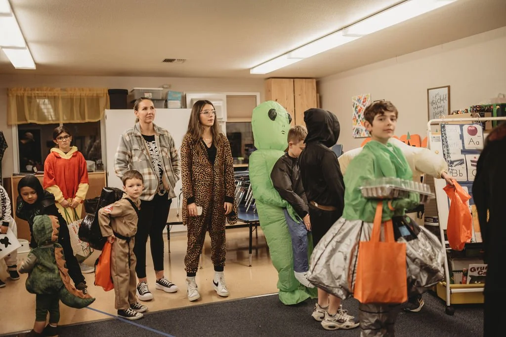 Children and adults dressed in costumes standing in a classroom or community room, waiting in line.