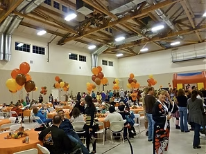 People gathered in a decorated indoor hall with orange and black balloons, sitting at tables and socializing, possibly for a celebration or event.
