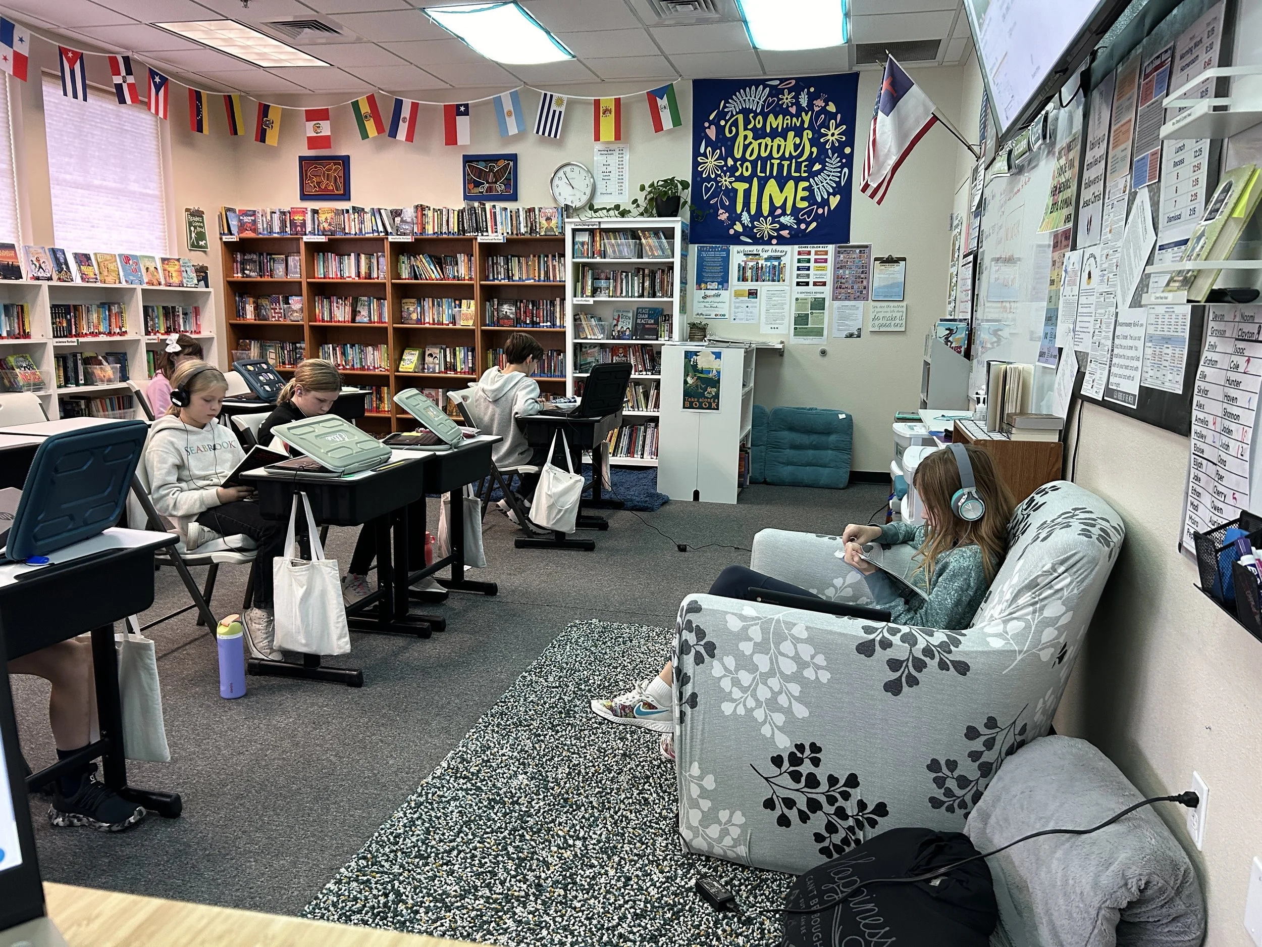 Children in a library classroom sitting at desks with laptops, listening with headphones; one girl is sitting on a patterned armchair reading a device, surrounded by bookshelves, and posters on the wall.