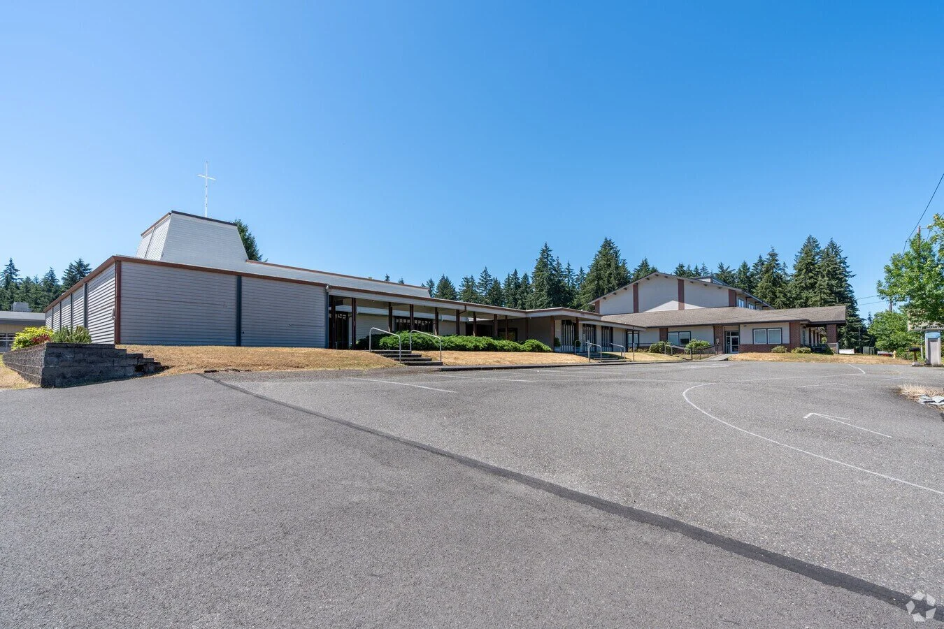 A church building with a cross on top, situated on a hill with a parking lot in the foreground, surrounded by trees under a clear blue sky.