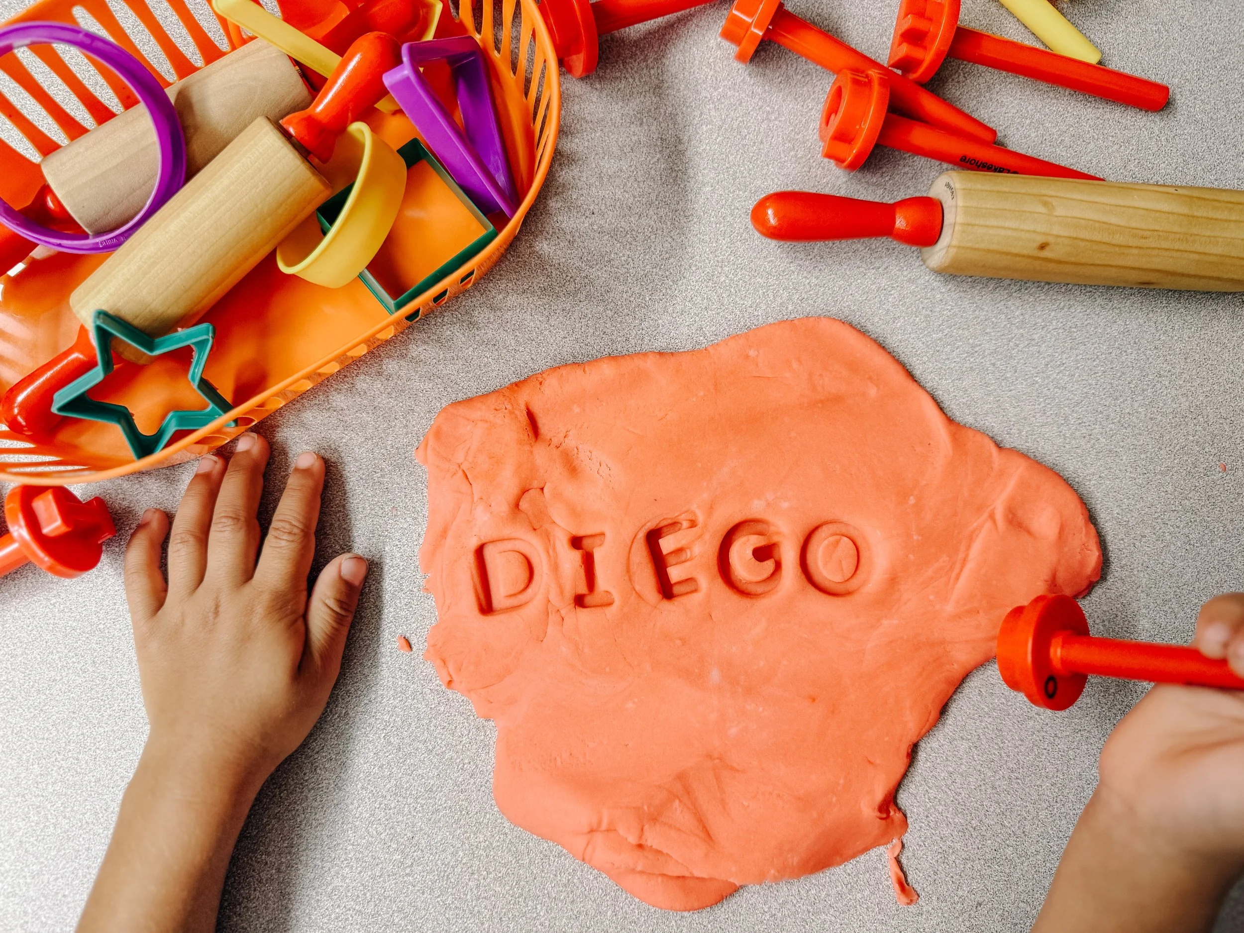 Child's hand pressing cookie cutter with the letters "DIEGO" into orange playdough on a table. A basket of playdough tools is nearby.