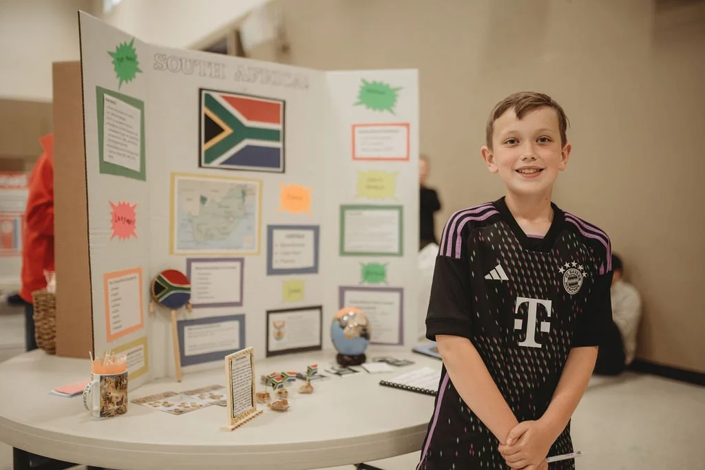 A smiling boy standing in front of a South Africa-themed school project display board, which includes the South African flag and related decorations.