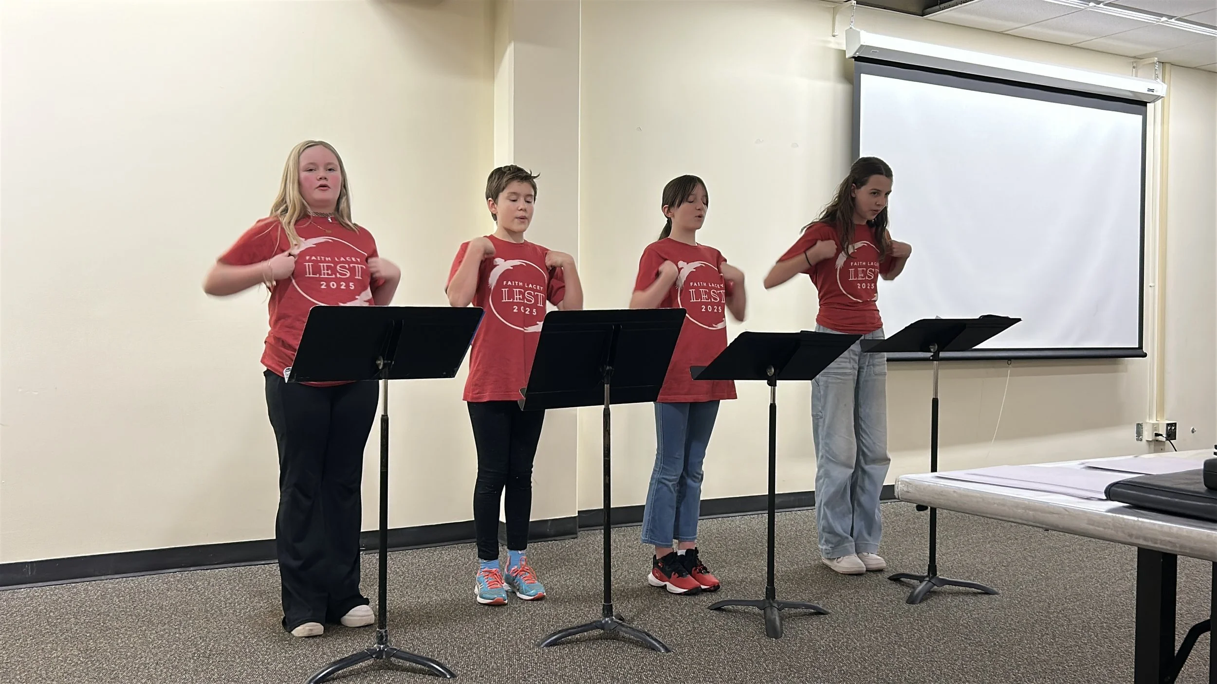 Four girls standing in a line in front of music stands, singing or reciting, with a large white projection screen behind them, all wearing matching red T-shirts with a white graphic and text, in a room with beige walls and gray carpet.