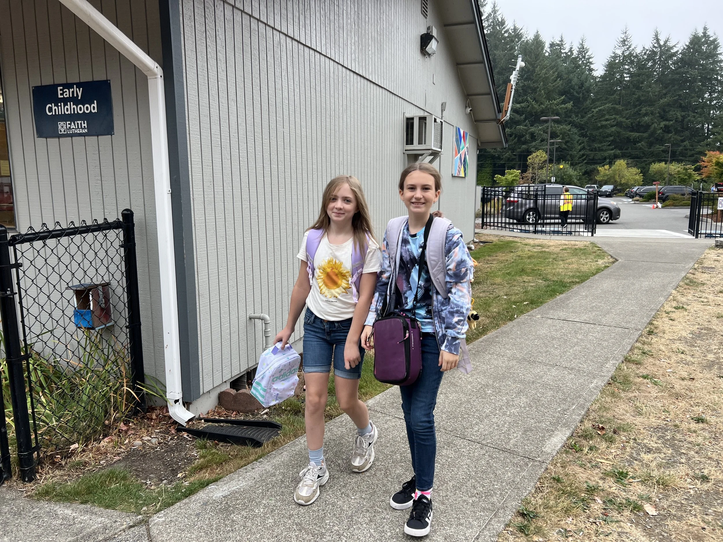 Two young girls walking outside a church building. One girl with a purple backpack and floral jacket, the other with a yellow sunflower t-shirt, holding a backpack. The church sign reads 'Early Childhood Faith Lutheran.' Gated parking lot and a person in yellow vest are visible in the background.