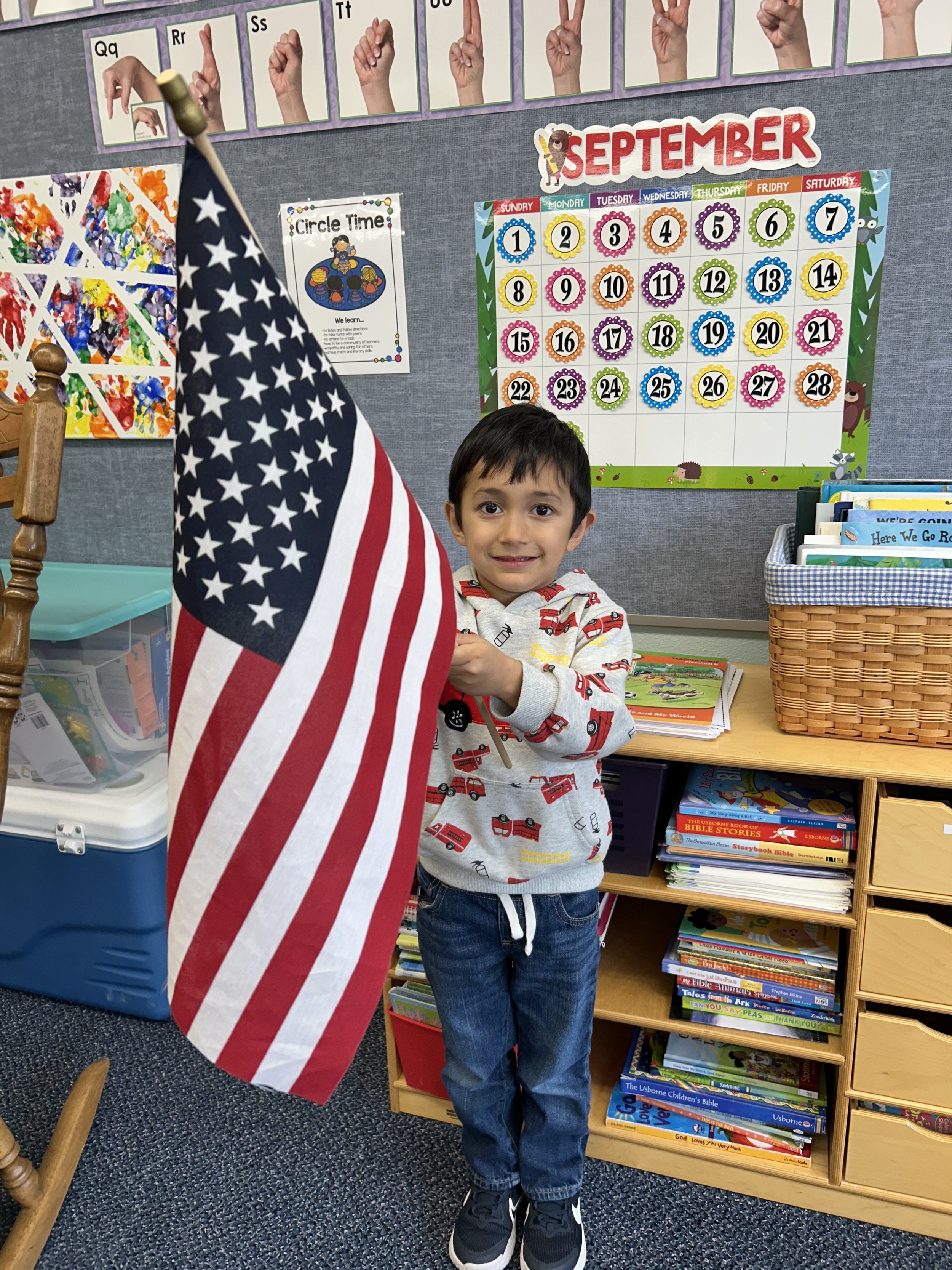 A young boy standing in a classroom holding an American flag, with a colorful calendar and educational posters on the bulletin board behind him.