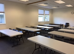 Empty classroom with white tables, black chairs, beige walls, and two windows
