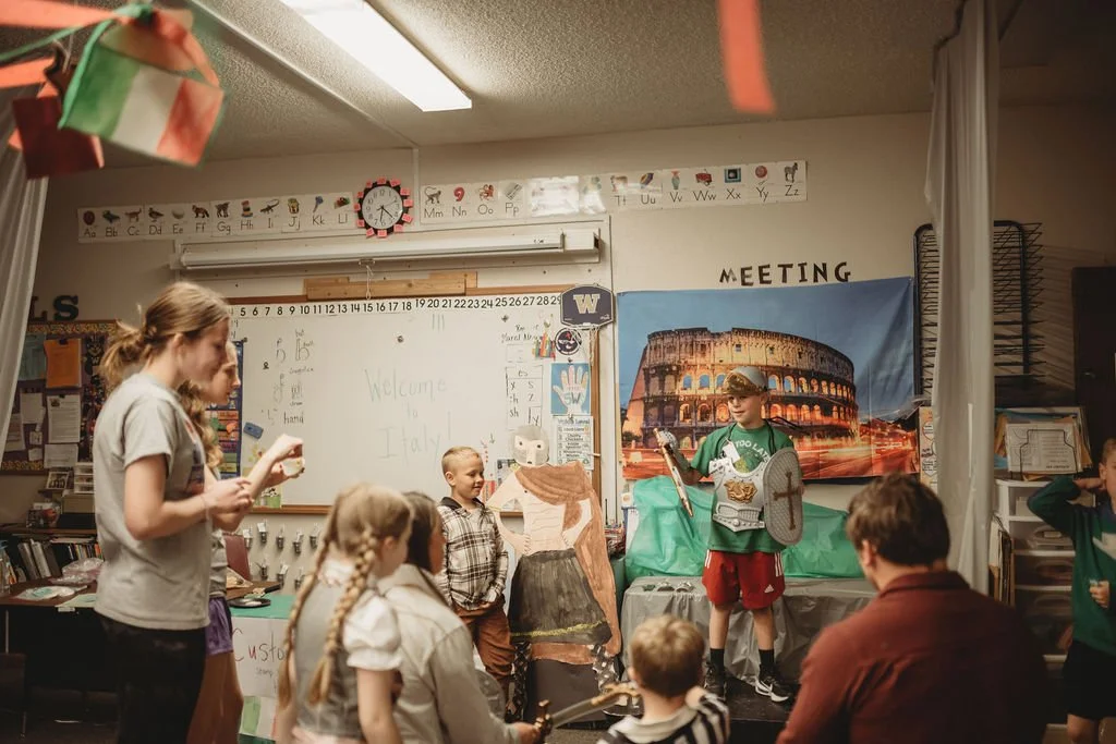 Children dressed as medieval knights and characters performing a play in a classroom decorated with a backdrop of the Colosseum in Rome.