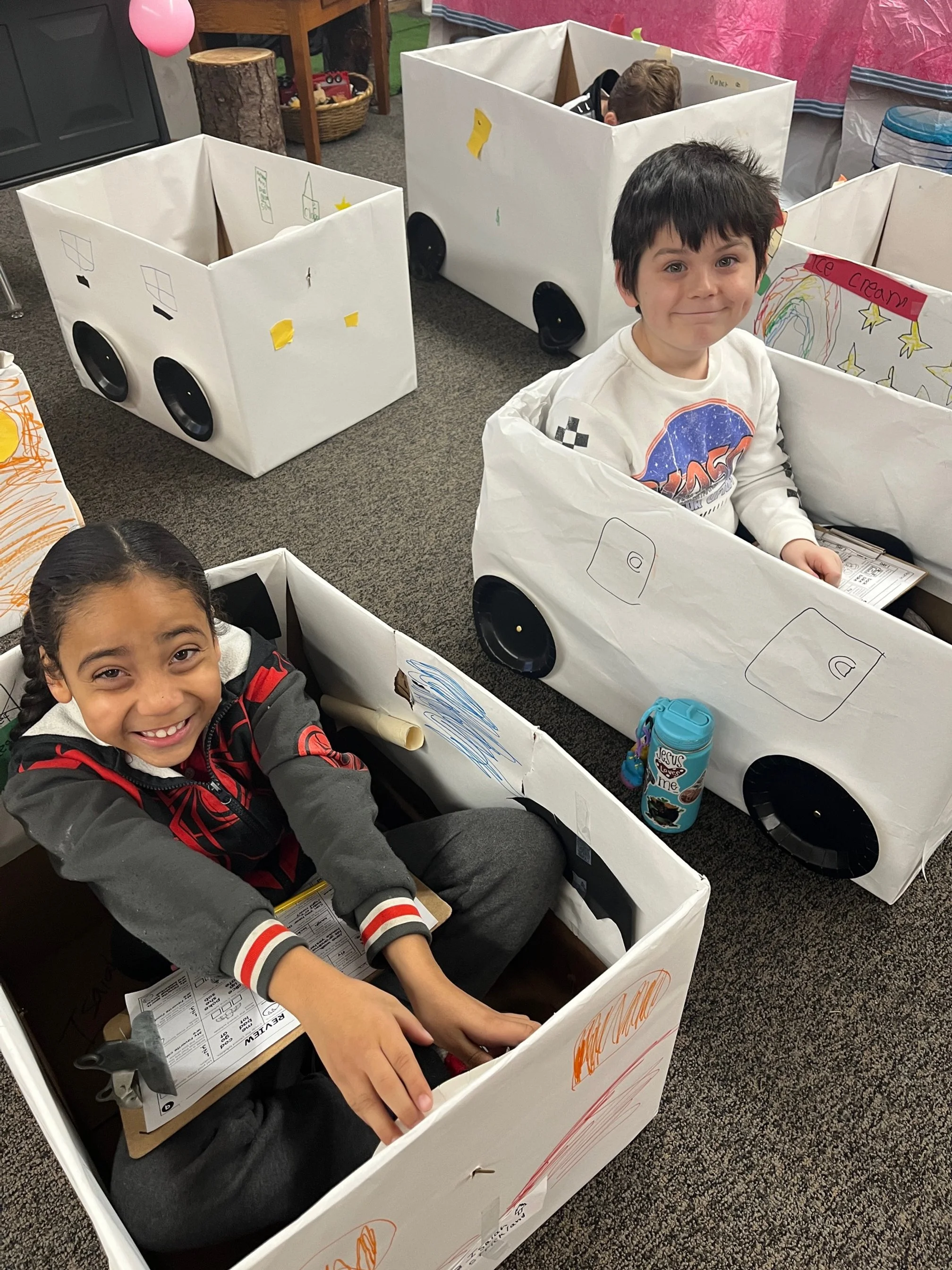 Two children sitting inside cardboard car-shaped boxes. The boxes are decorated to look like cars with wheels and drawings. The children are smiling and appear happy, with one girl in a gray and red jacket and one boy in a white shirt with dinosaur print.