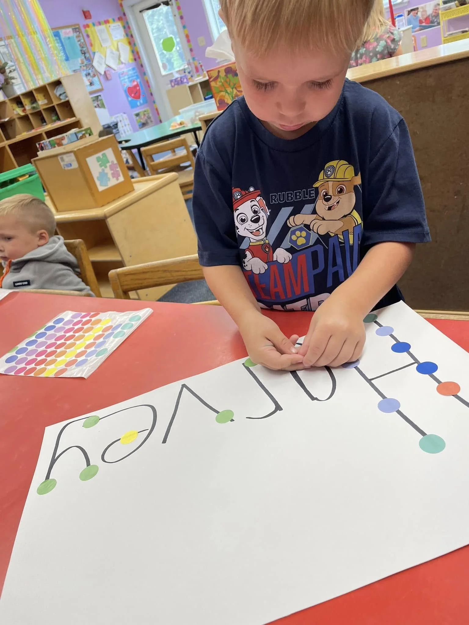 A young boy is placing colorful dots on a large white poster with the word 'birthday' written on it in a classroom. The classroom has colorful decorations and other children in the background.