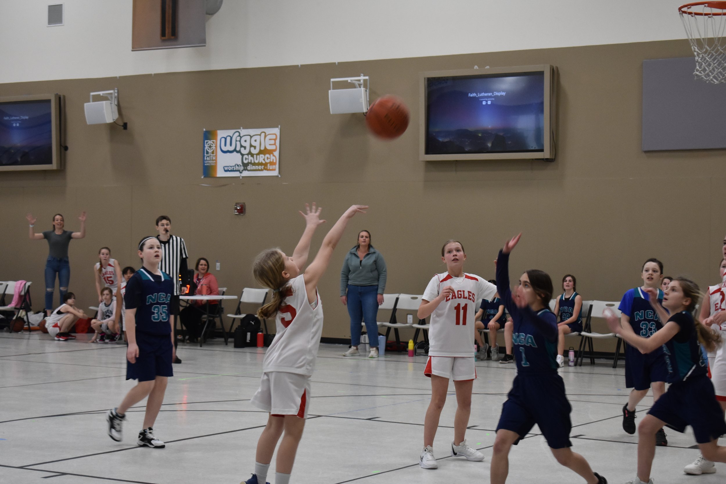 Children playing basketball in an indoor gym with spectators and a referee.