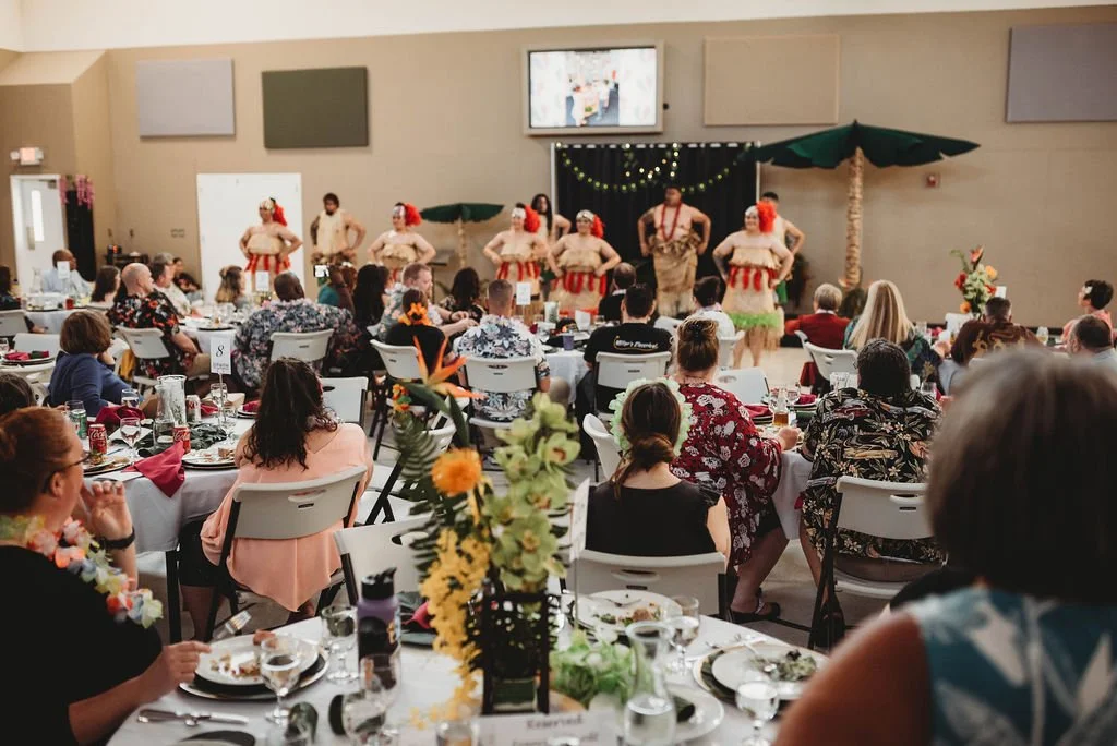 Hula dancers performing on stage at a banquet with guests seated at decorated tables.