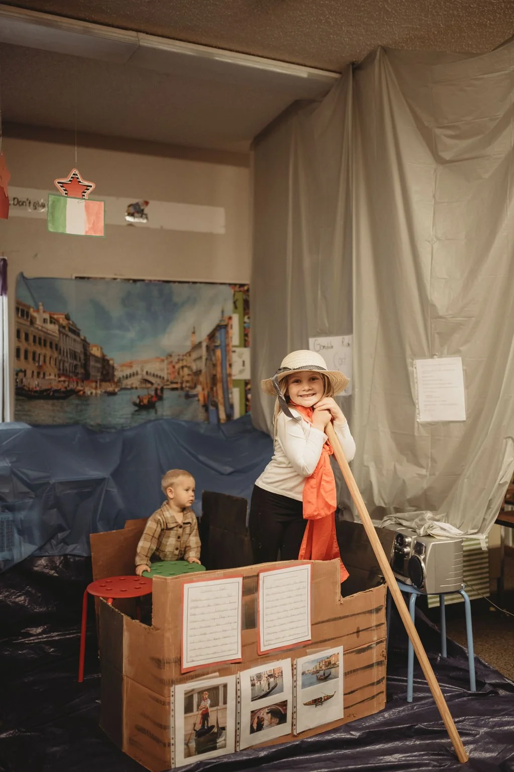 Children dressed in costumes participating in a school play or project, with a Venice, Italy themed background, a cardboard boat, and Luca and Marco photos on display.