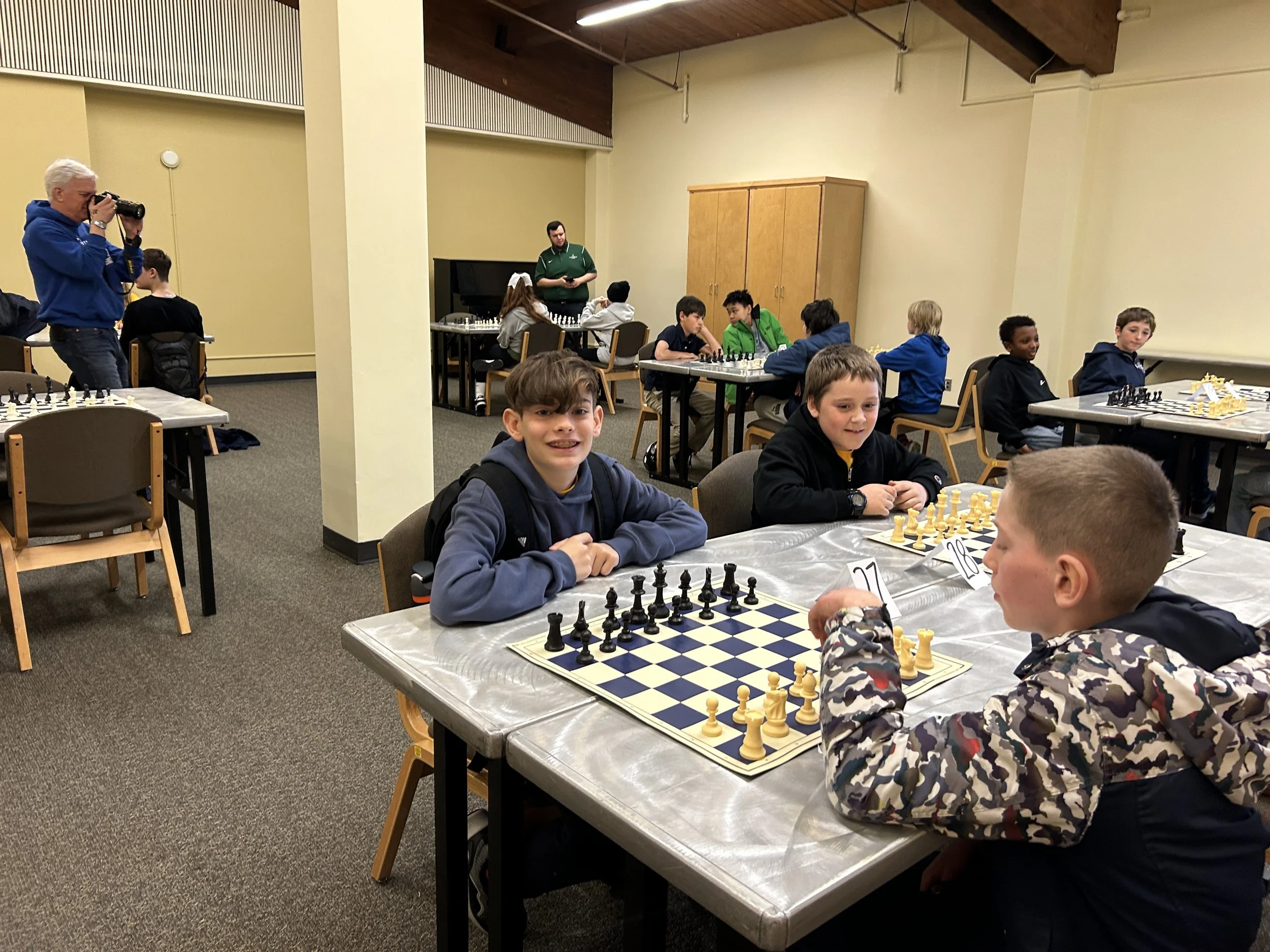 Young boys playing chess at a tournament, some looking at the camera and smiling, in a large room with other players and a photographer in the background.