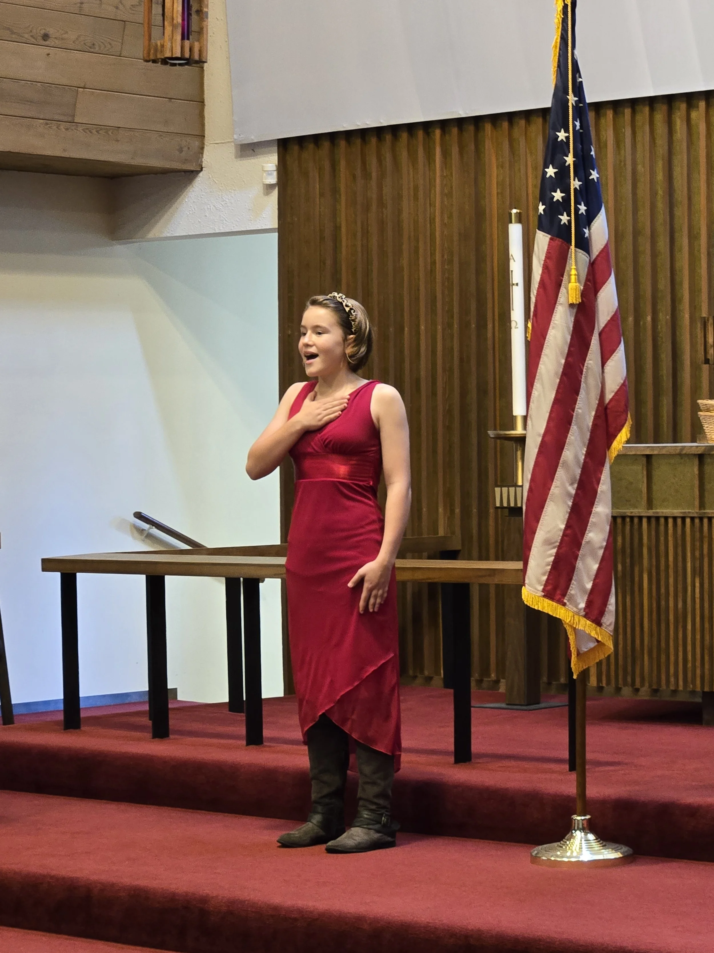 A girl in a red dress and black boots singing with her hand on her chest on a stage with a U.S. flag to her side.