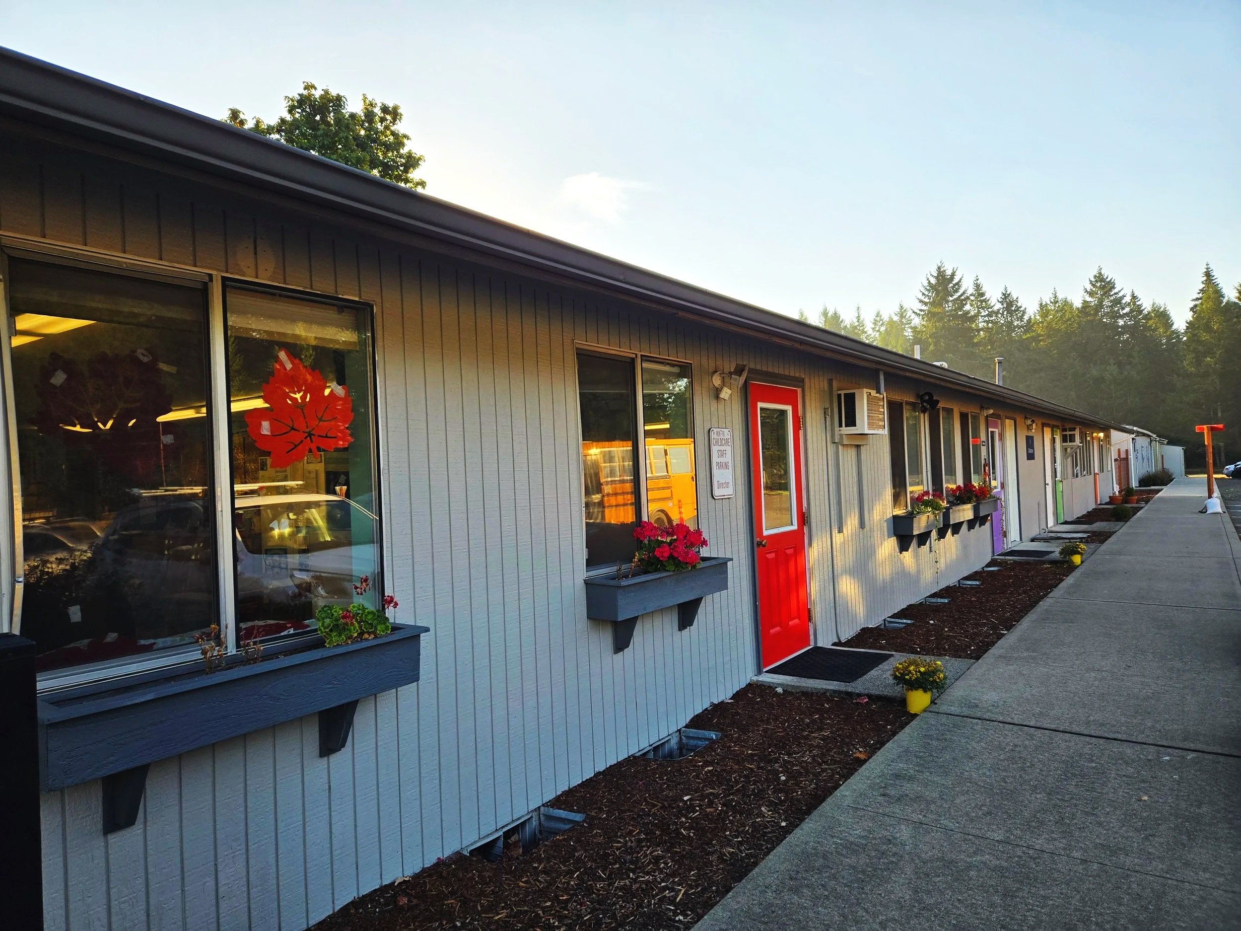 Exterior view of a single-story building with a gray siding, red door, window boxes with flowers, and an air conditioning unit, with a sidewalk and trees in the background.