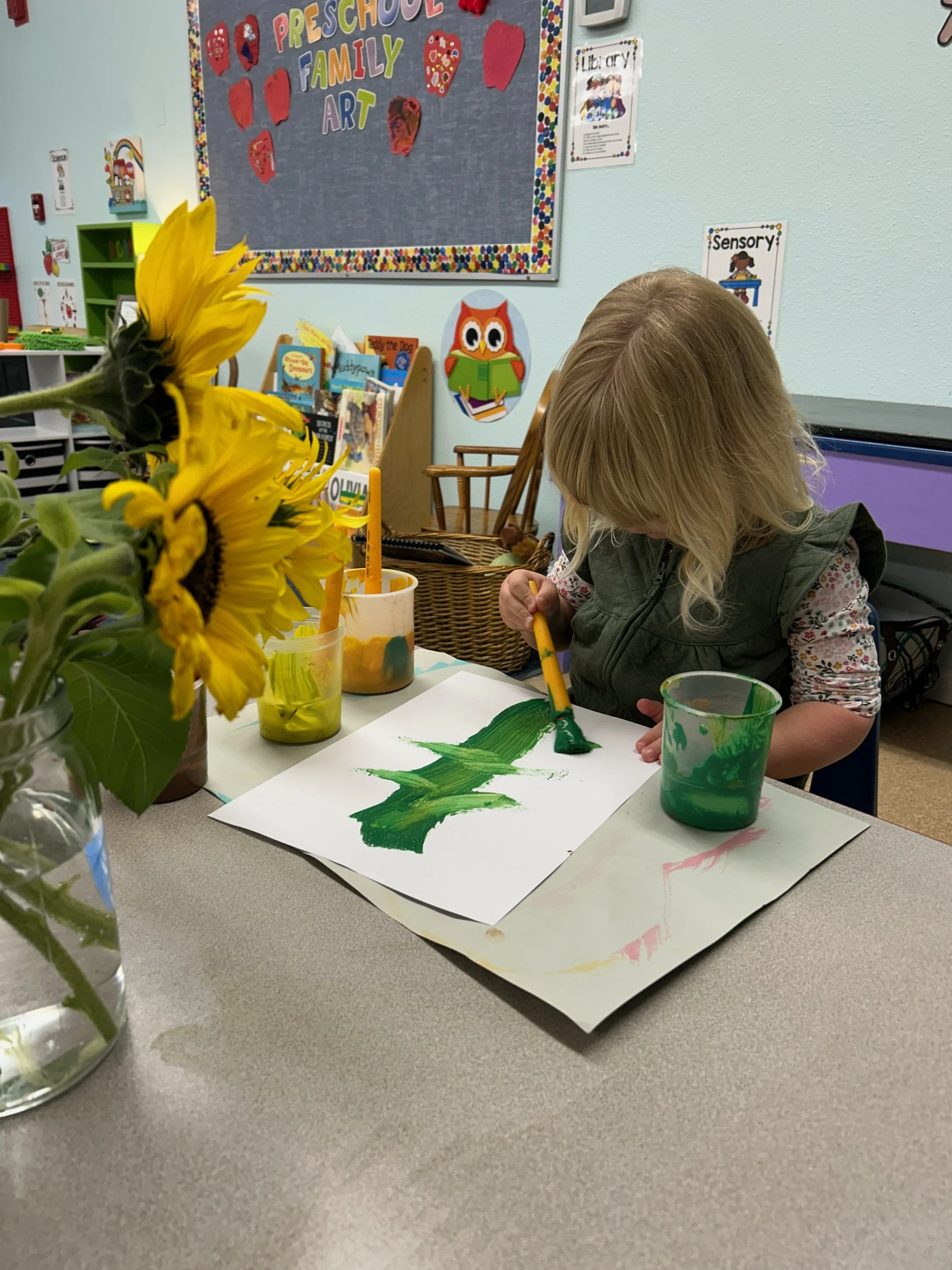 A young girl with blonde hair painting green on a sheet of paper in a preschool classroom, with sunflower flowers in a vase in the foreground. The classroom has educational posters, books, and art displays on the walls.