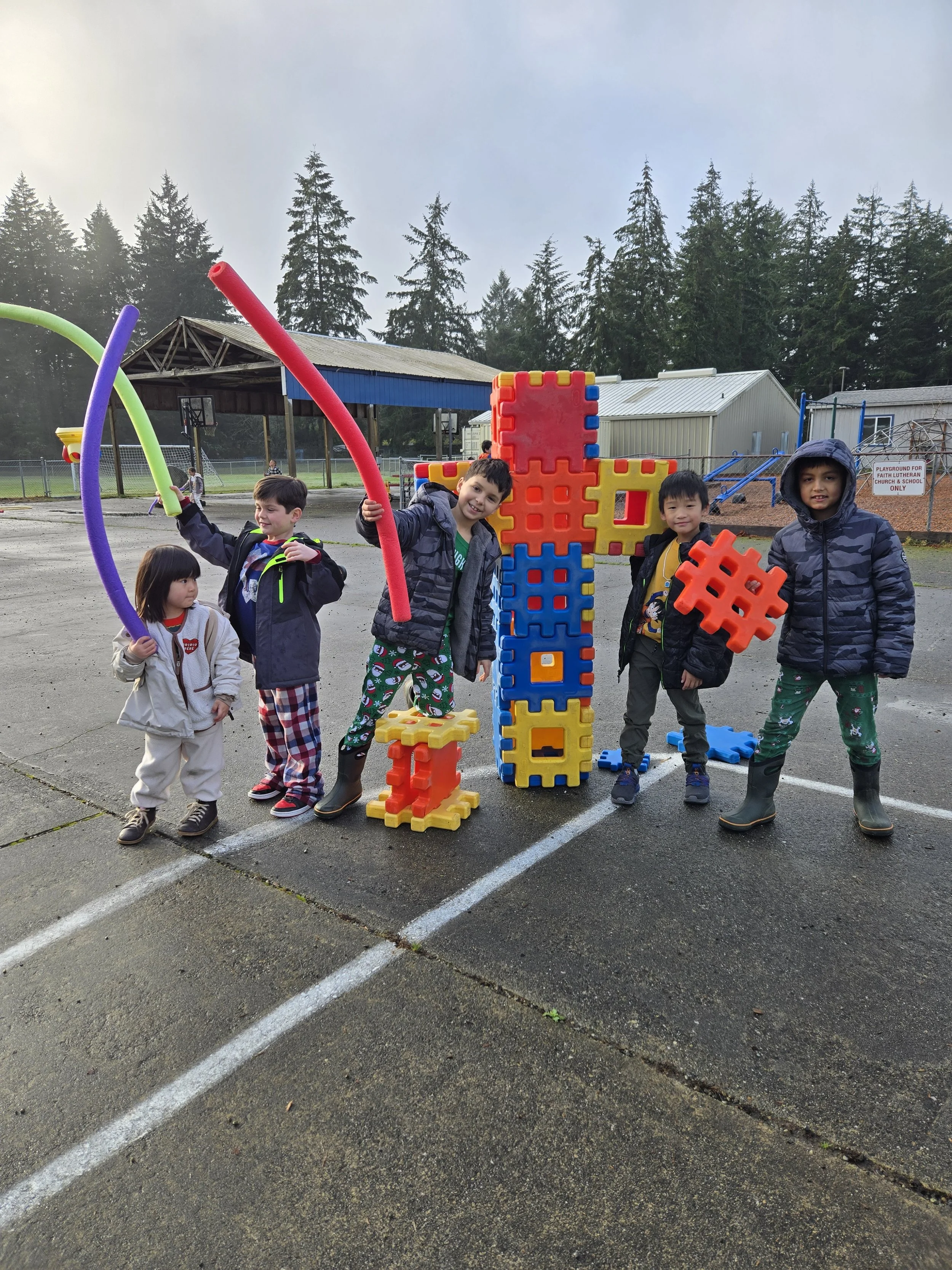 Children playing outdoors with large foam building blocks on a playground in a parking lot, surrounded by trees and buildings.