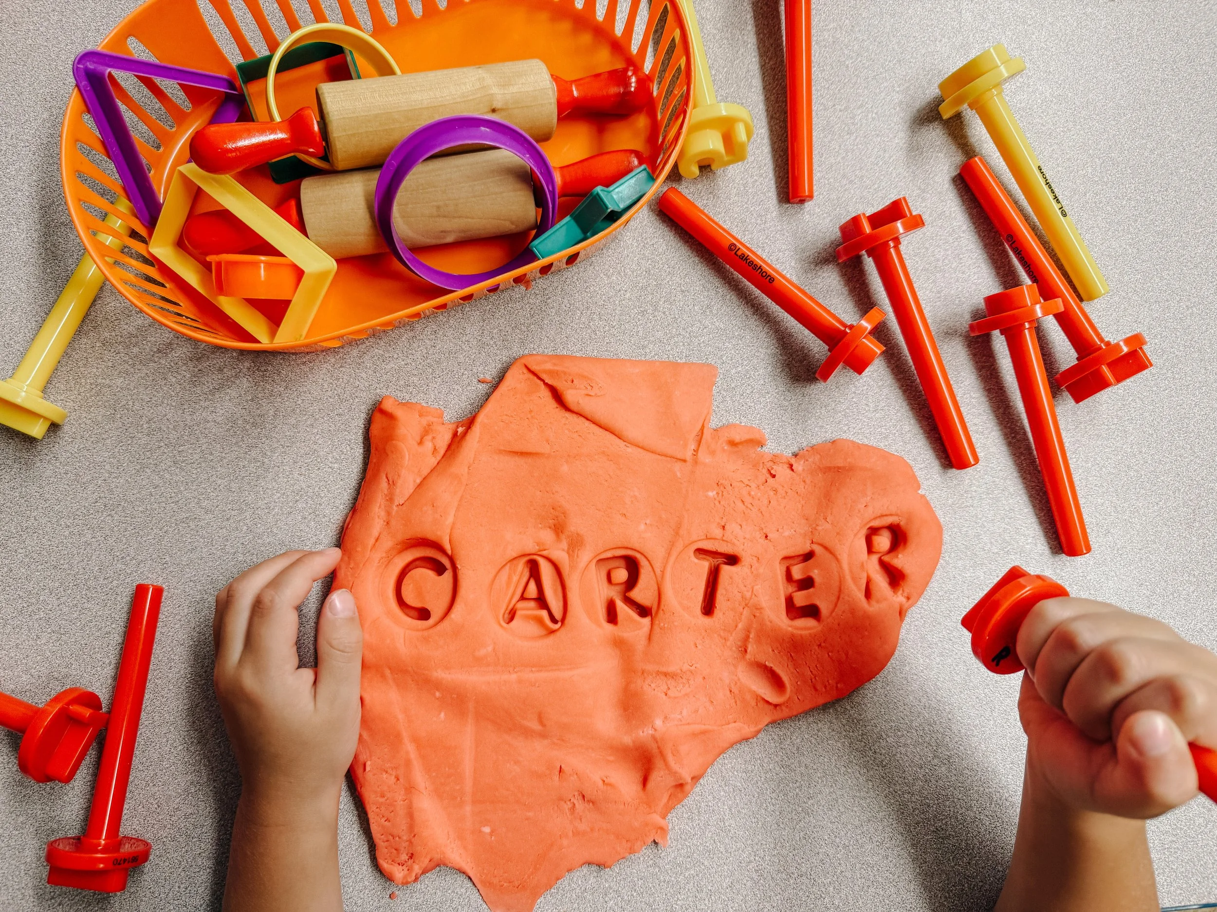 Child using cookie cutters to cut the word "CARTER" into orange playdough on a table. There are various plastic tools, cookie cutters, and a basket of playdough tools and shapes nearby.
