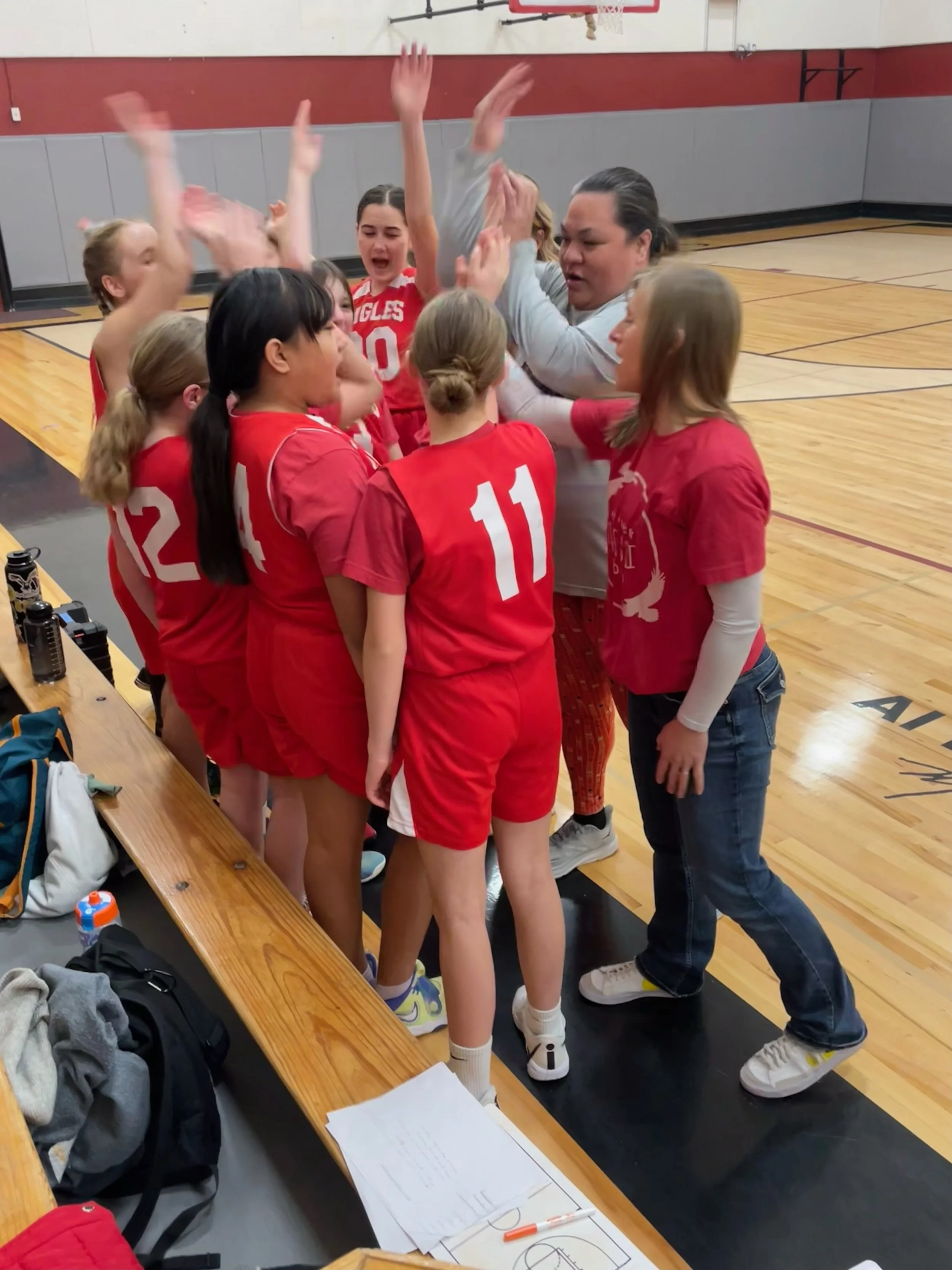 A girls' volleyball team huddles with their coach on a basketball court, celebrating during a game or practice.