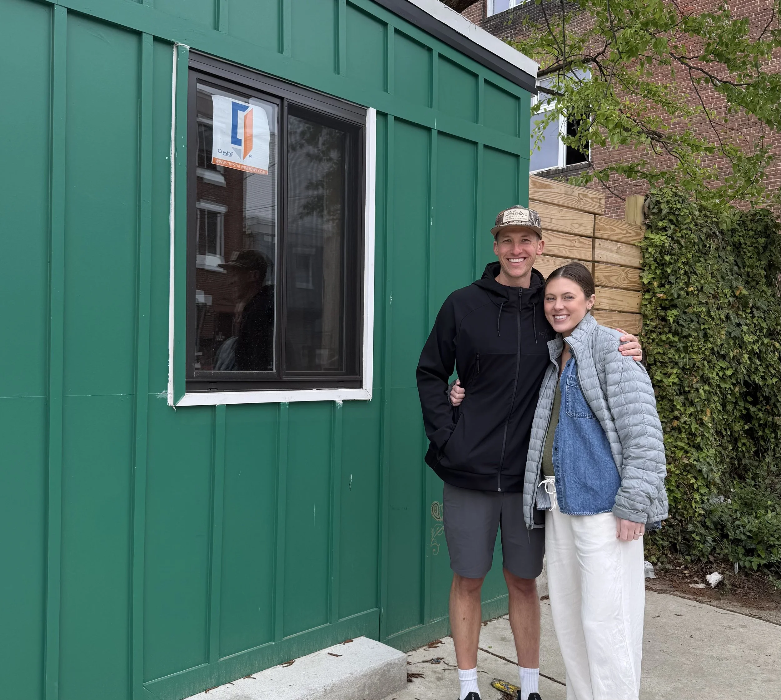 A man and a woman smiling and standing close together outdoors in front of a green building with a window and a brick building in the background. The man is wearing a black jacket, shorts, two-tone socks, and a cap. The woman is wearing a light gray jacket, a blue shirt, and white pants.