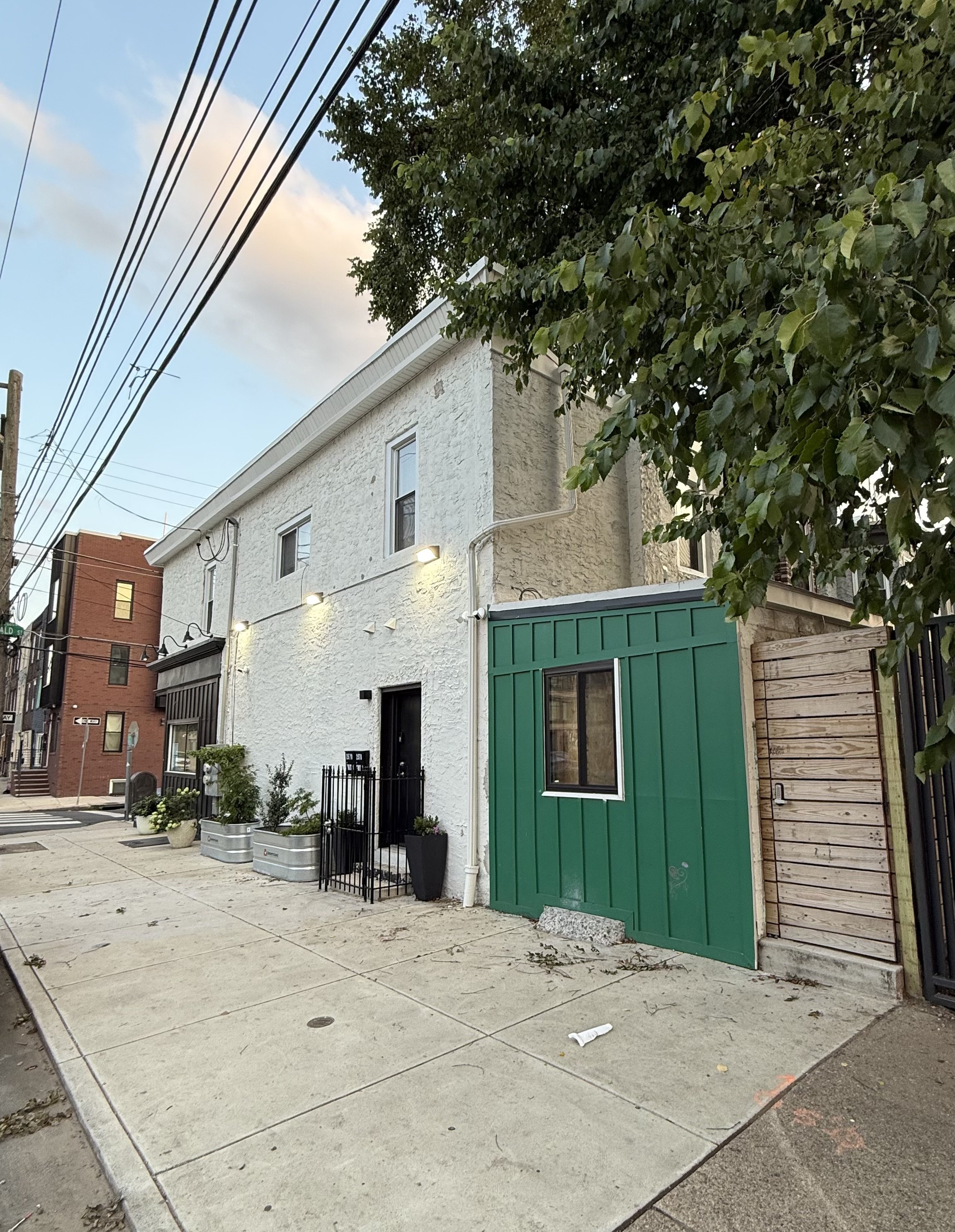 View of a building with white textured walls, green storefront, and black sidewalk patio fencing. Potted plants and small trees are in front. Overhead power lines and leafy tree partially obscure the building. The street corner sign shows the intersection with a brick building and other city structures in the background.