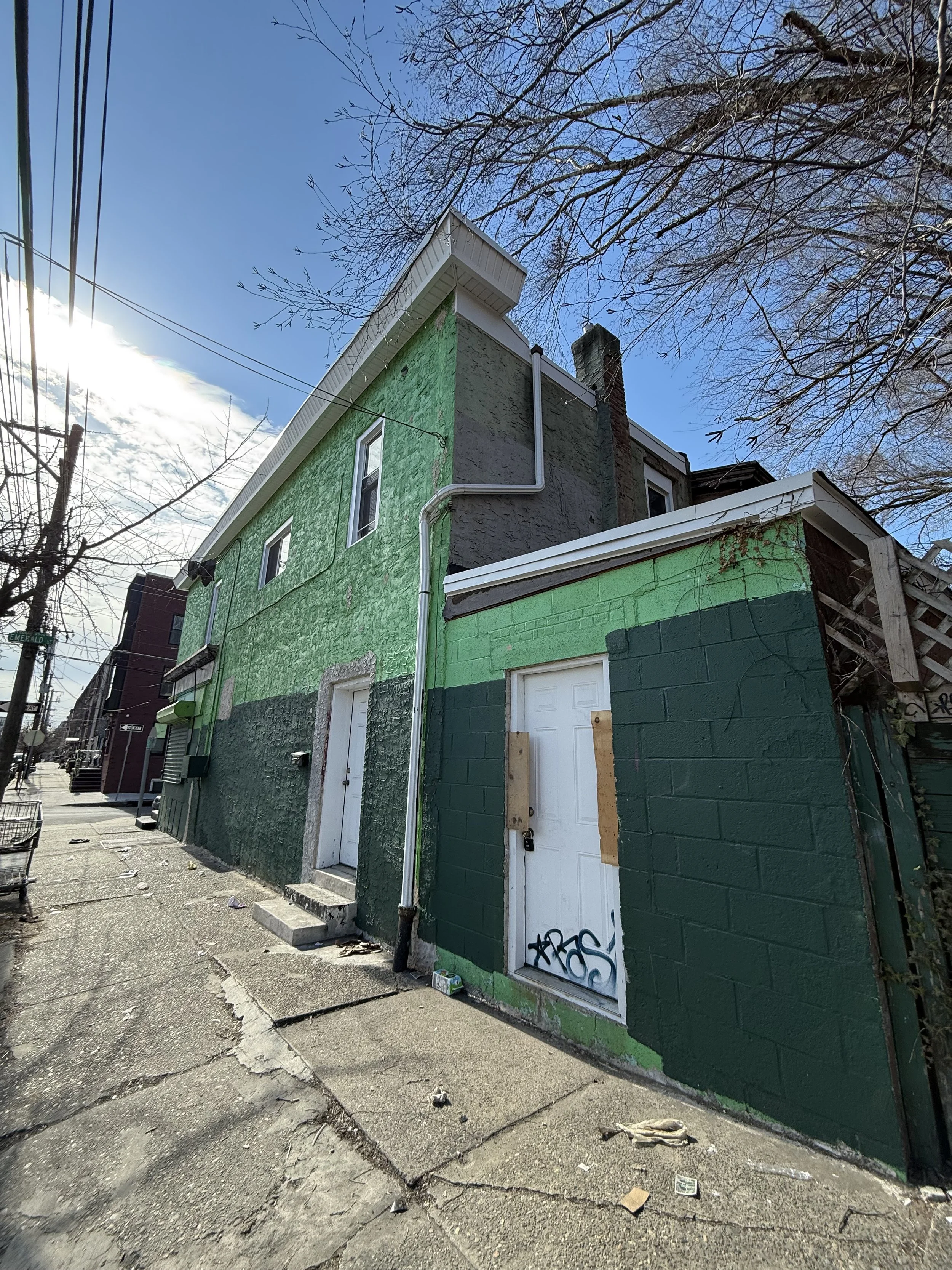A green painted building with two garage doors and a white door on an urban sidewalk, with leafless trees and a partly cloudy sky overhead.